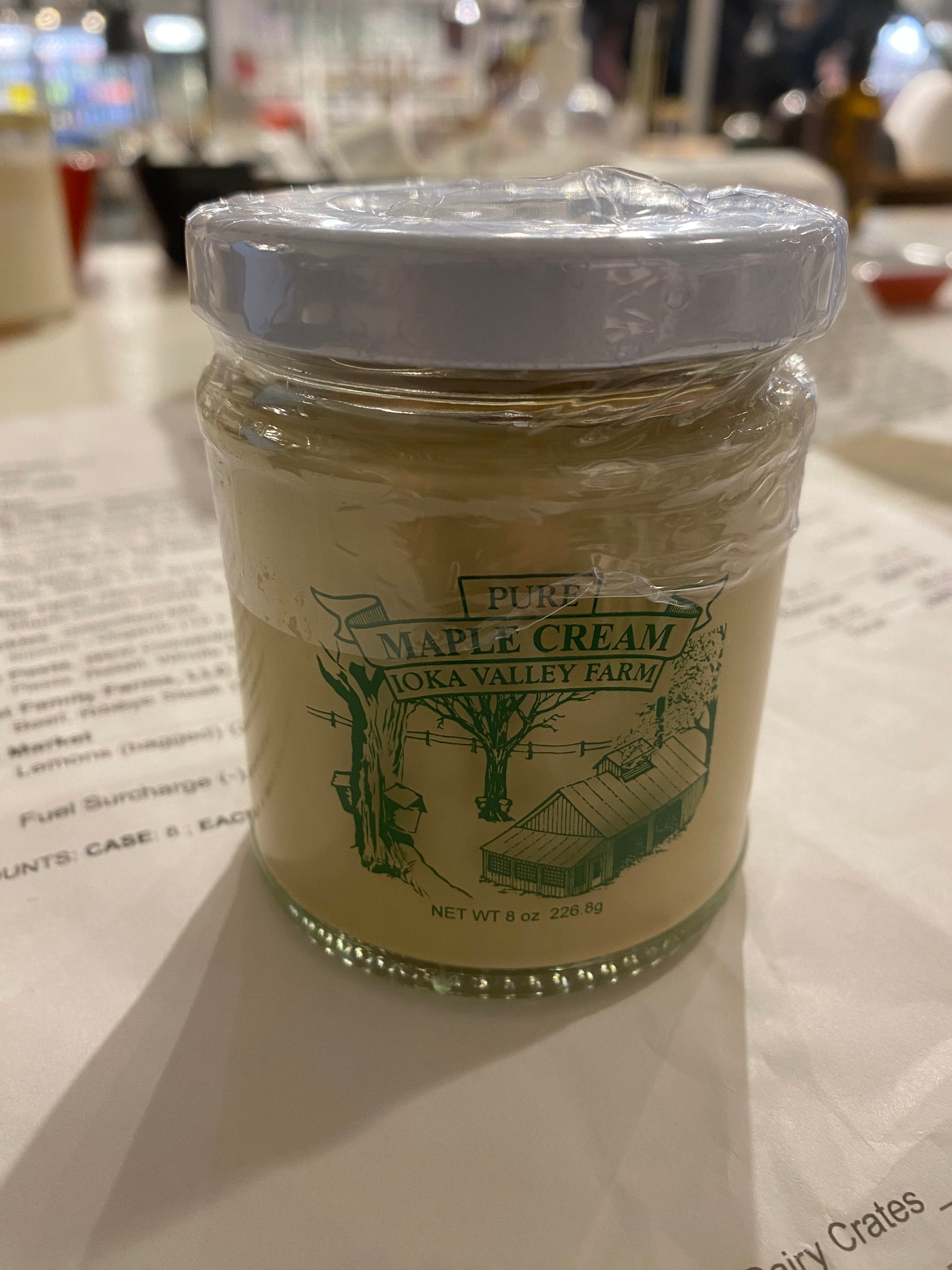 A jar of Westerlind Maple Cream from Oka Valley Farm, sealed with a white cap, displayed on a table with blurred background.
