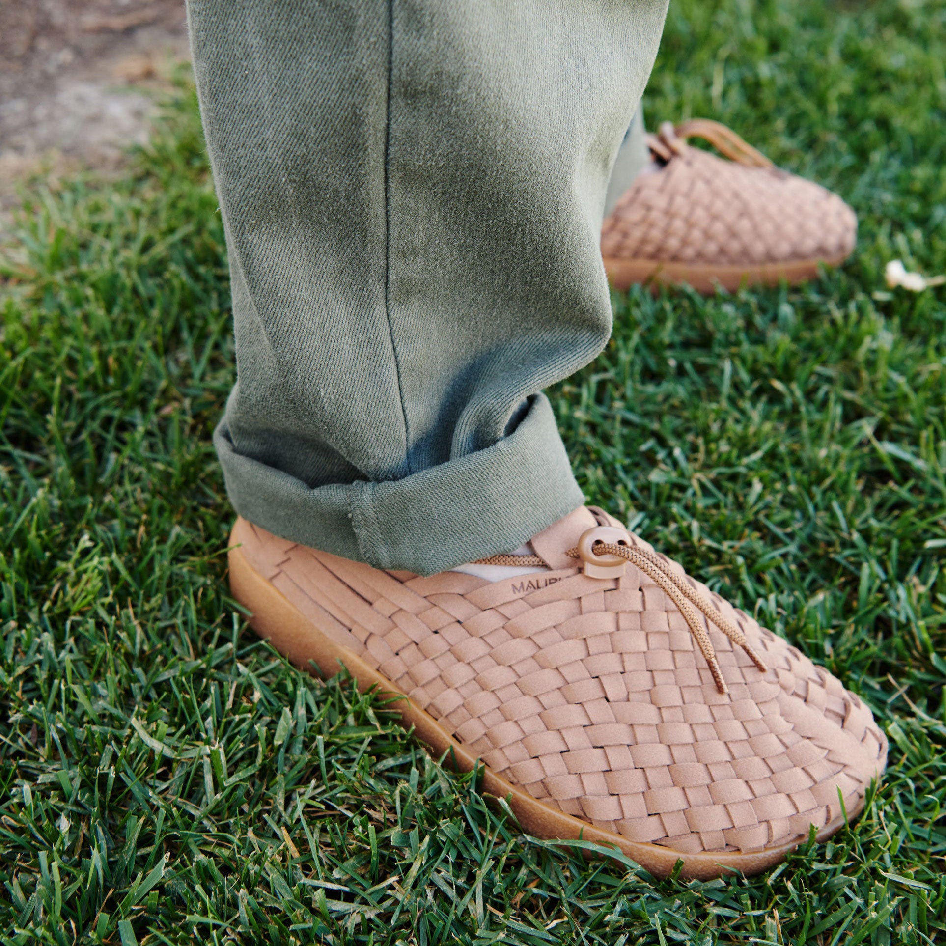 A person wearing Malibu Sandals M Latigo light brown vegan leather slip-ons and green pants stands on grass.
