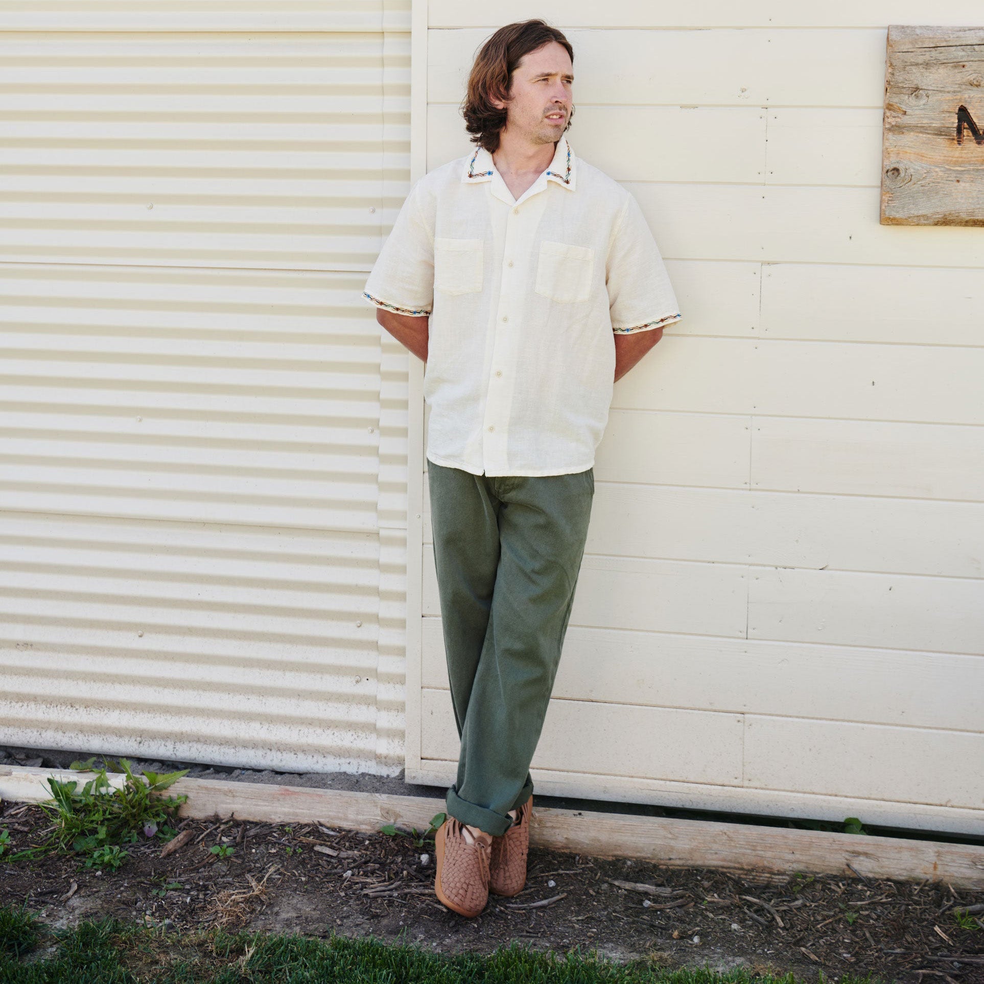 A man stands outside against a white wall with his hands behind his back, wearing a short-sleeve white shirt, green pants, and brown M Latigo shoes by Malibu Sandals crafted from vegan leather.