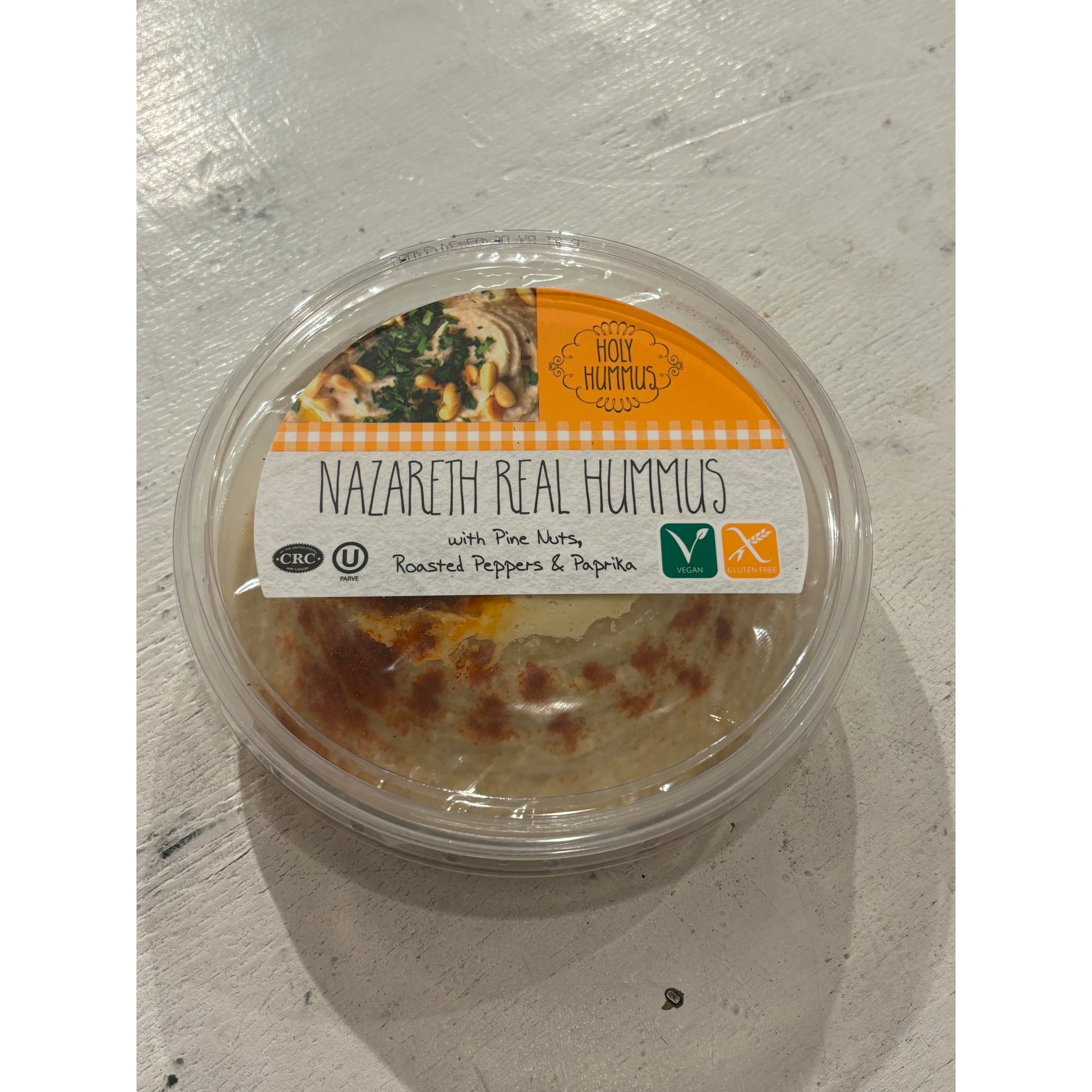 Clear plastic container of "Hummus Nazareth - Holy Hummus" with roasted peppers and pine nuts, displayed on a white wooden surface.