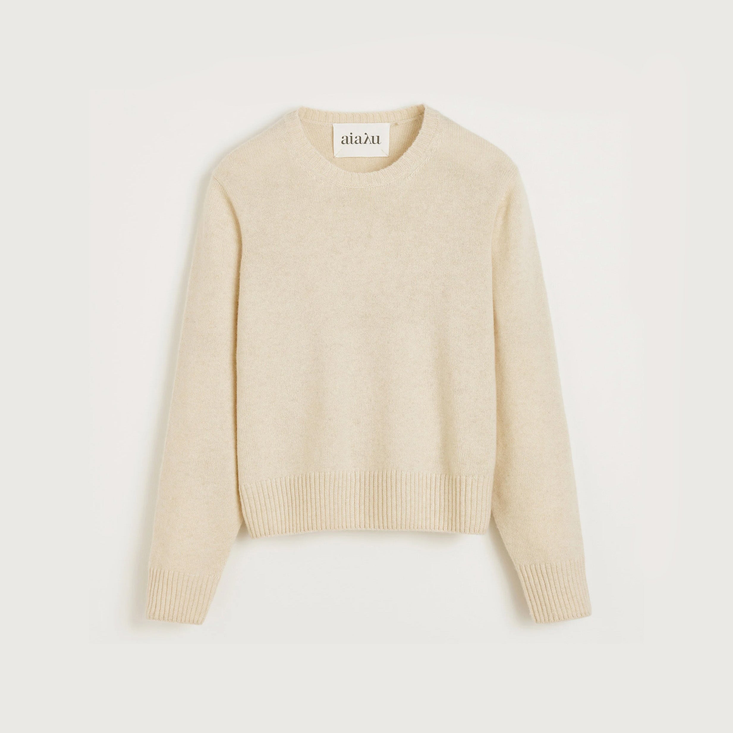 The Aiayu Inga Sweater is a beige, long-sleeve cashmere piece with classic ribbed cuffs and hem, elegantly showcased against a neutral backdrop.