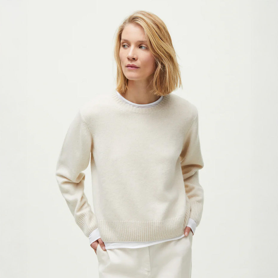 A person with blond hair wears the Inga Sweater by Aiayu, a smooth-knitted cream cashmere piece, paired with white pants. They stand against a plain light background, gaze turned to the side, hands in pockets.
