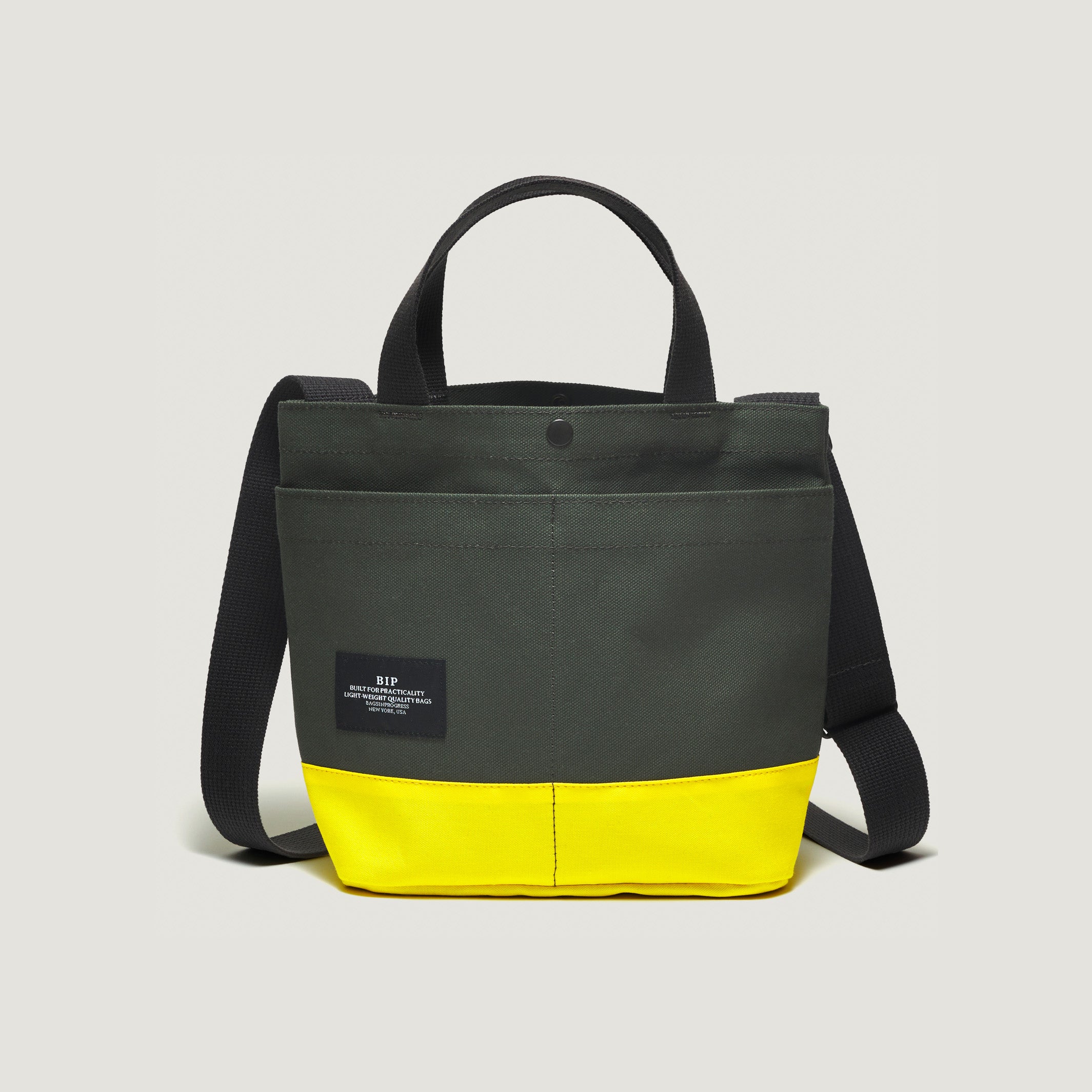 The New York Tote Small by Bags in Progress, Special Edition: Forest Green/Yellow, features black handles and a shoulder strap. It includes an external pocket for added functionality and has a black label with white text on the front.