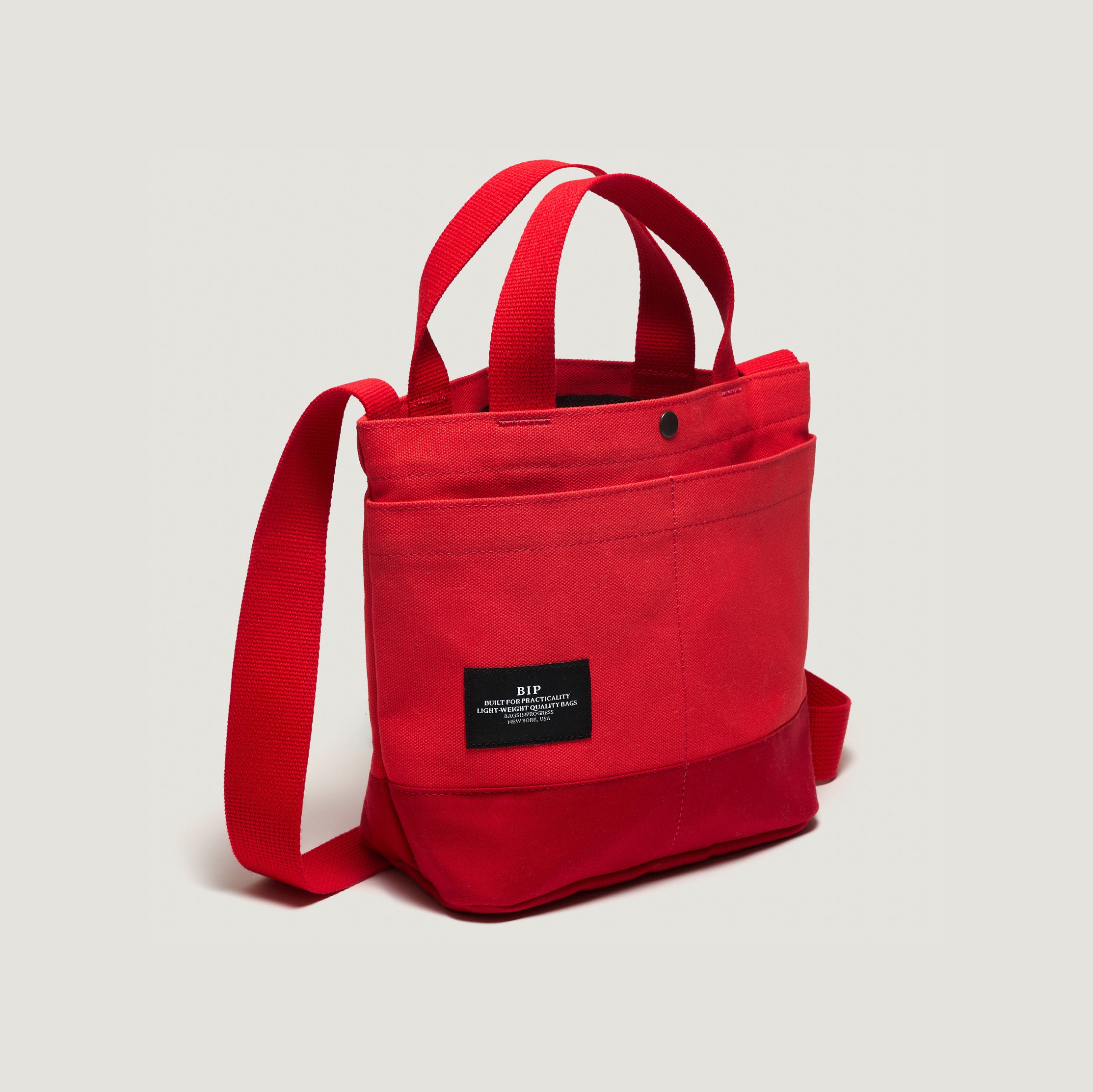 The New York Tote Small, Special Edition: Red/Red by Bags in Progress is a vibrant red canvas tote with dual handles and a handy shoulder strap. It features practical front pockets and a sleek black label with white text, effortlessly blending style and functionality.