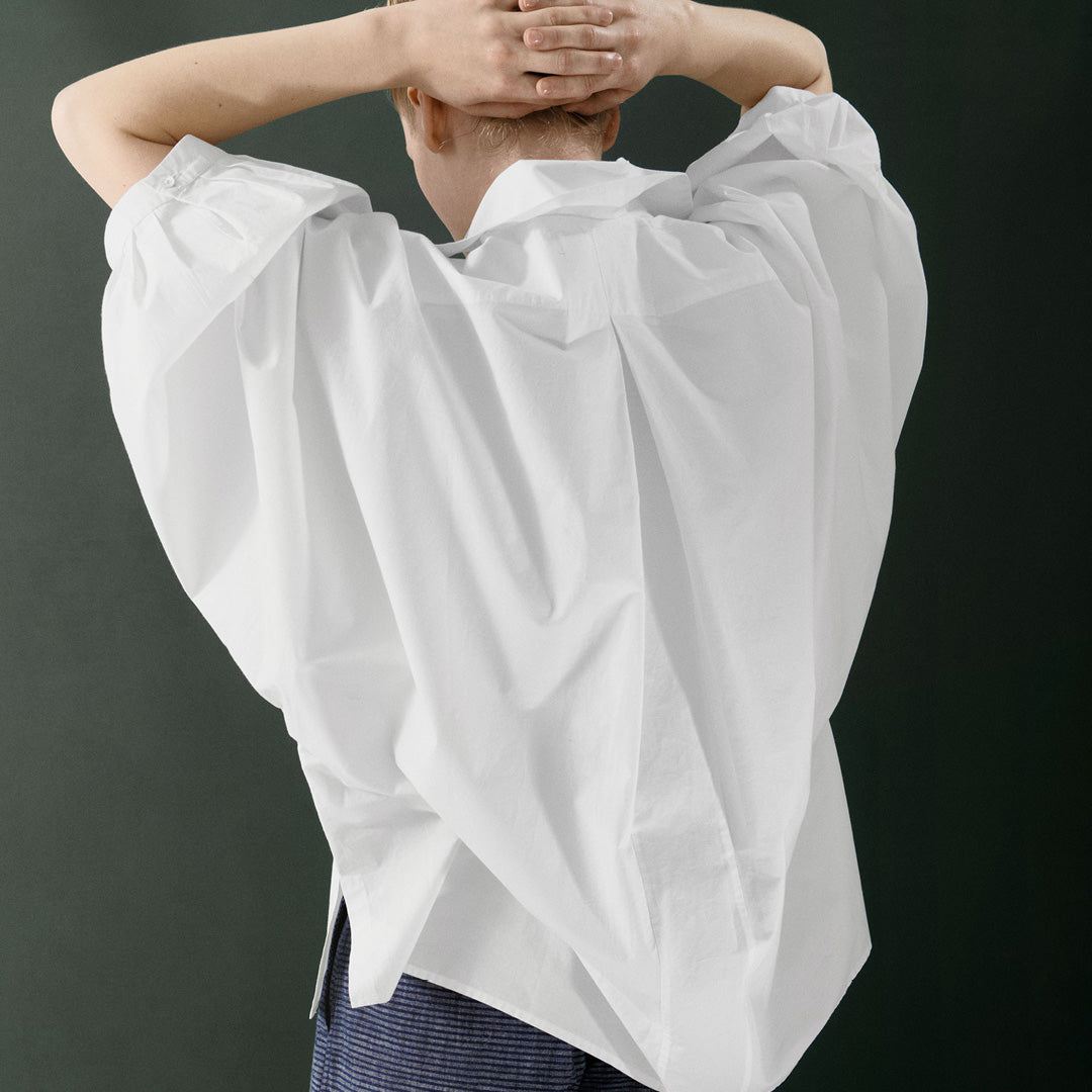 Wearing the Jan Machenhauer Nina Shirt in Poplin White, a person with their back to the camera stands in blue pants, arms raised and hands clasped behind their head.