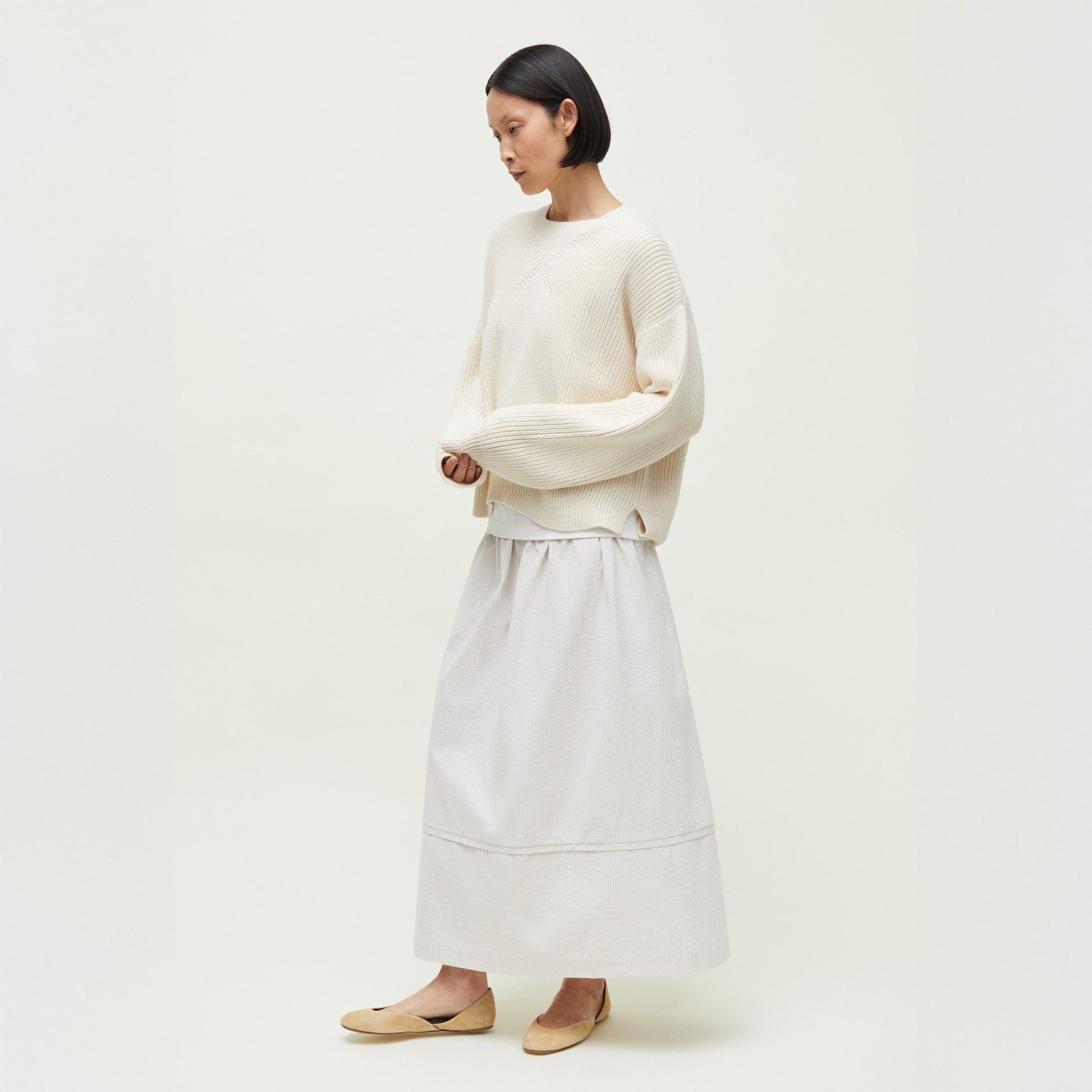 A woman stands against a plain background wearing the Aiayu Kindra Sweater, an organic cotton cream knit, paired with a long white skirt and beige flats.