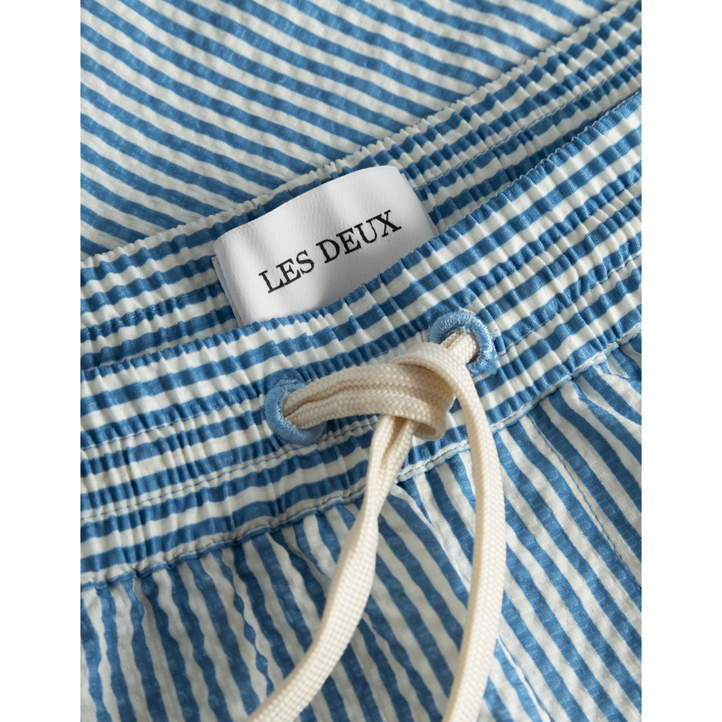 Close-up of a Stan Stripe Seersucker Swim Shorts in Washed Denim Blue/Light Ivory with a "Les Deux" label and a white drawstring.