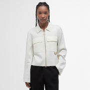 A woman stands against a white background wearing the Barbour Alma Knitted Zip Through Cardigan, crafted from a cotton-wool blend with large patch pockets, paired with black pants.