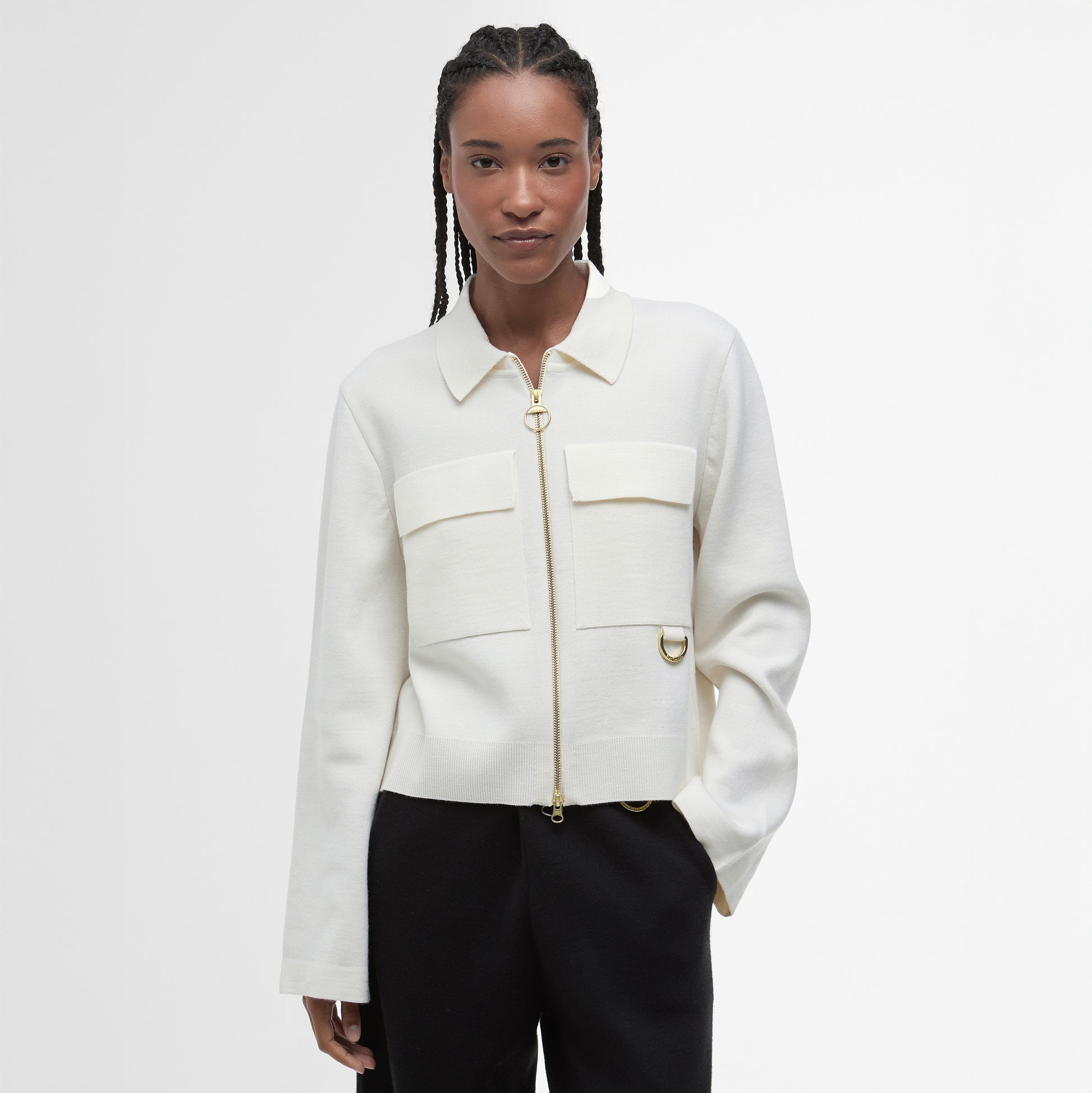 A woman stands against a white background wearing the Barbour Alma Knitted Zip Through Cardigan, crafted from a cotton-wool blend with large patch pockets, paired with black pants.