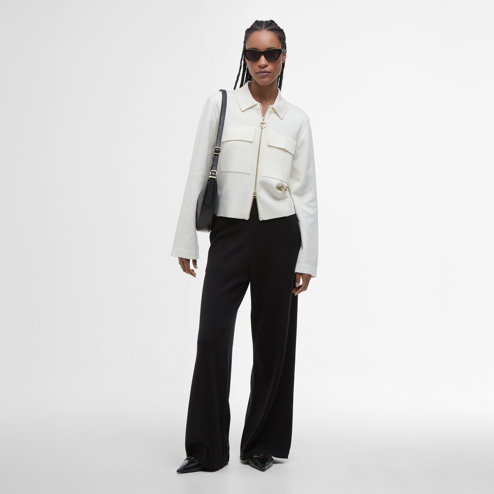 A person wearing a Barbour Alma Knitted Zip Through Cardigan with patch pockets, black wide-leg pants, pointed shoes, sunglasses, and a black shoulder bag stands against a plain white background.