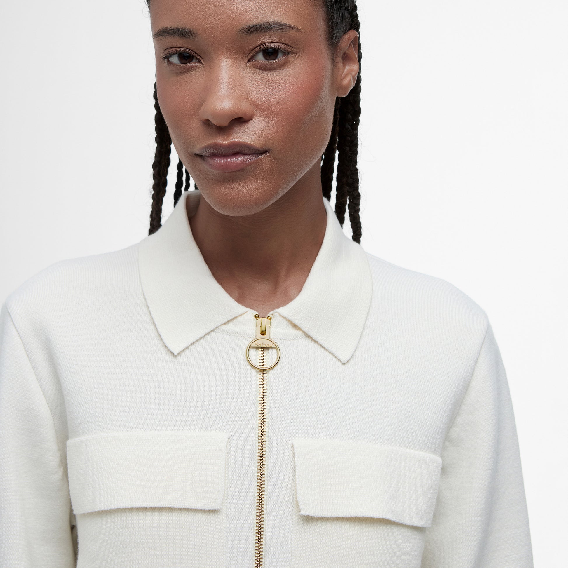 A person stands against a plain white background wearing Barbour's Alma Knitted Zip Through Cardigan in cream, crafted from a cotton-wool blend with a gold zipper and two patch pockets.