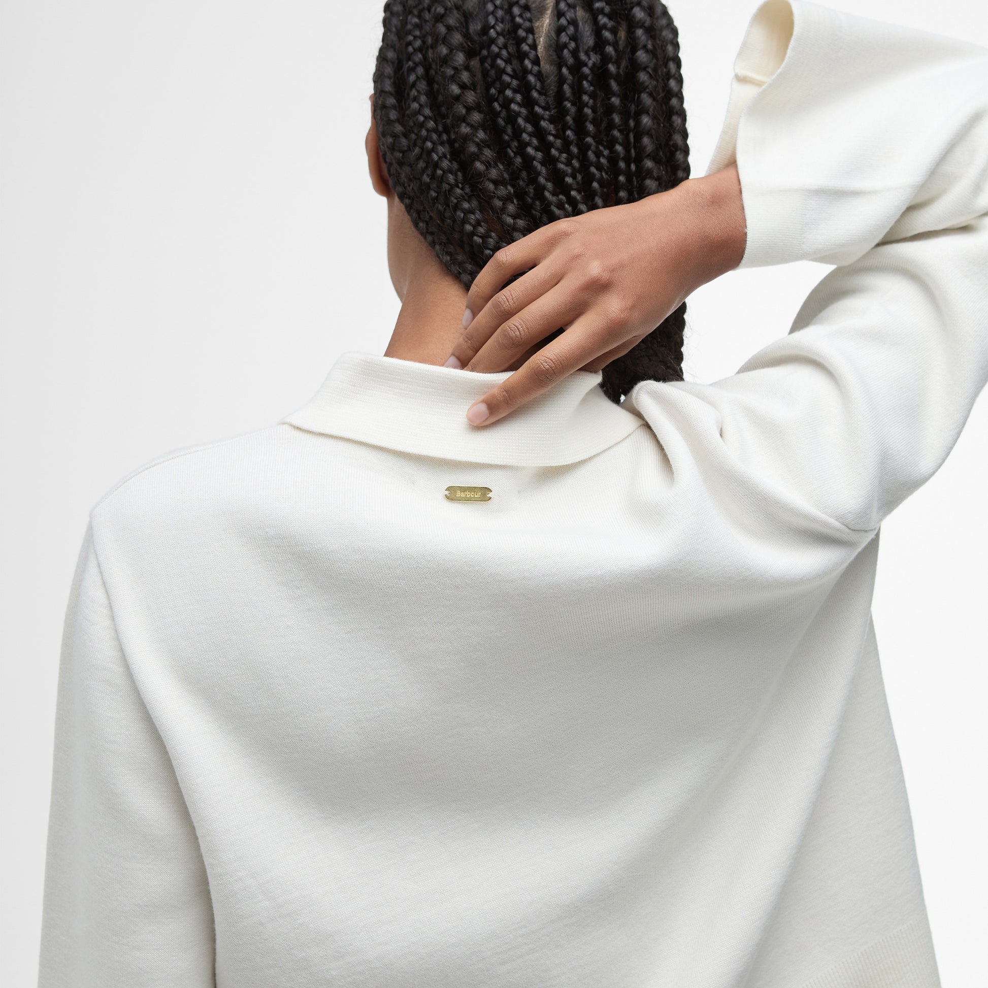 Person with braids, seen from behind, wearing the Barbour Alma Knitted Zip Through Cardigan—an off-white collared top crafted in a soft cotton wool blend—and touching the back of their neck.