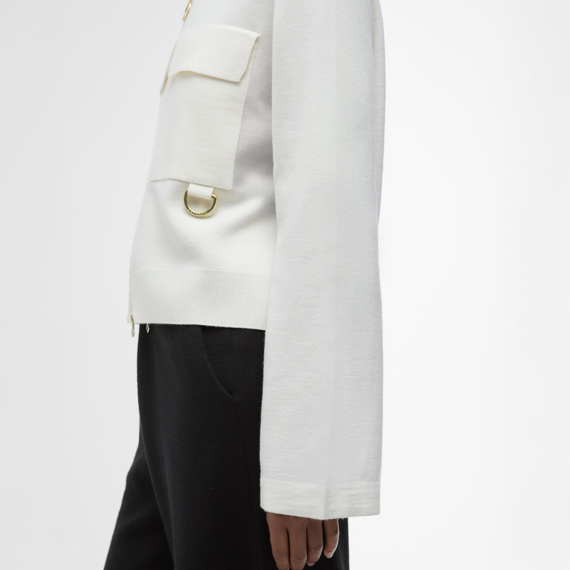 A person shown from the side against a plain background wears black pants and a Barbour Alma Knitted Zip Through Cardigan featuring gold hardware and patch pockets.