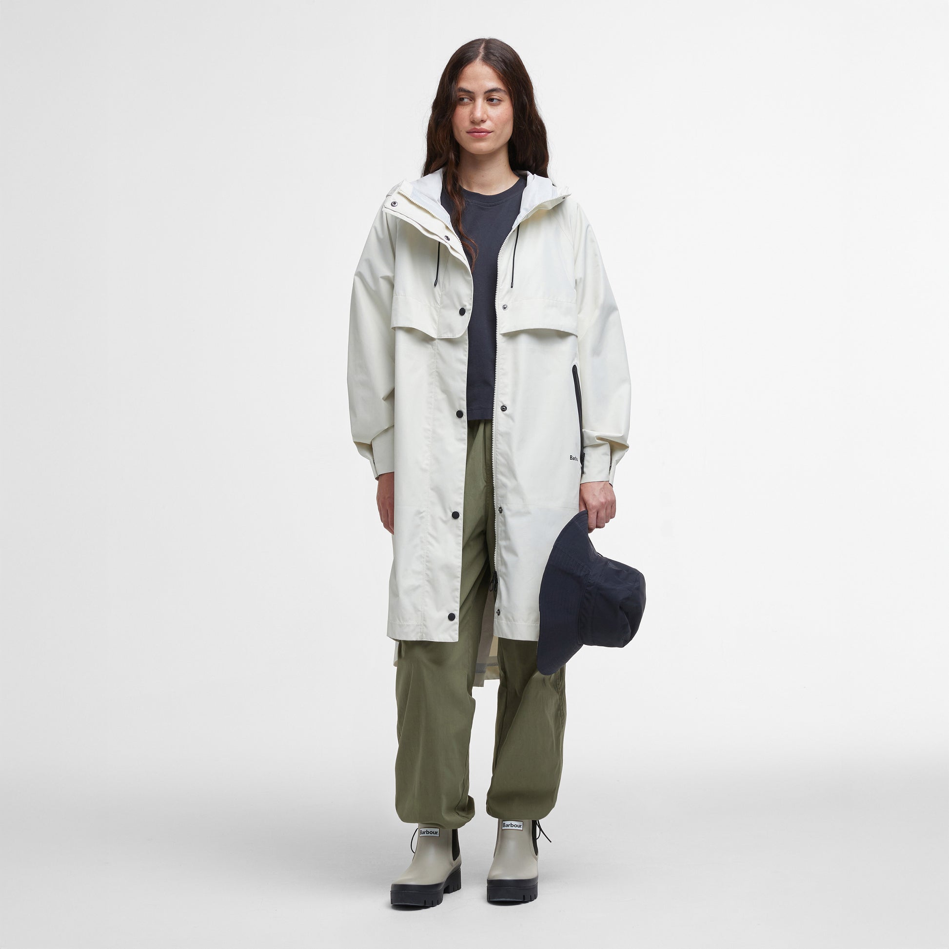 A person stands facing forward in a Barbour Jayla Waterproof Parka—long, white, and paired with a dark shirt, olive green pants, light boots, and holding a dark hat. Perfect for outdoor ventures. Shown against a plain white background.
