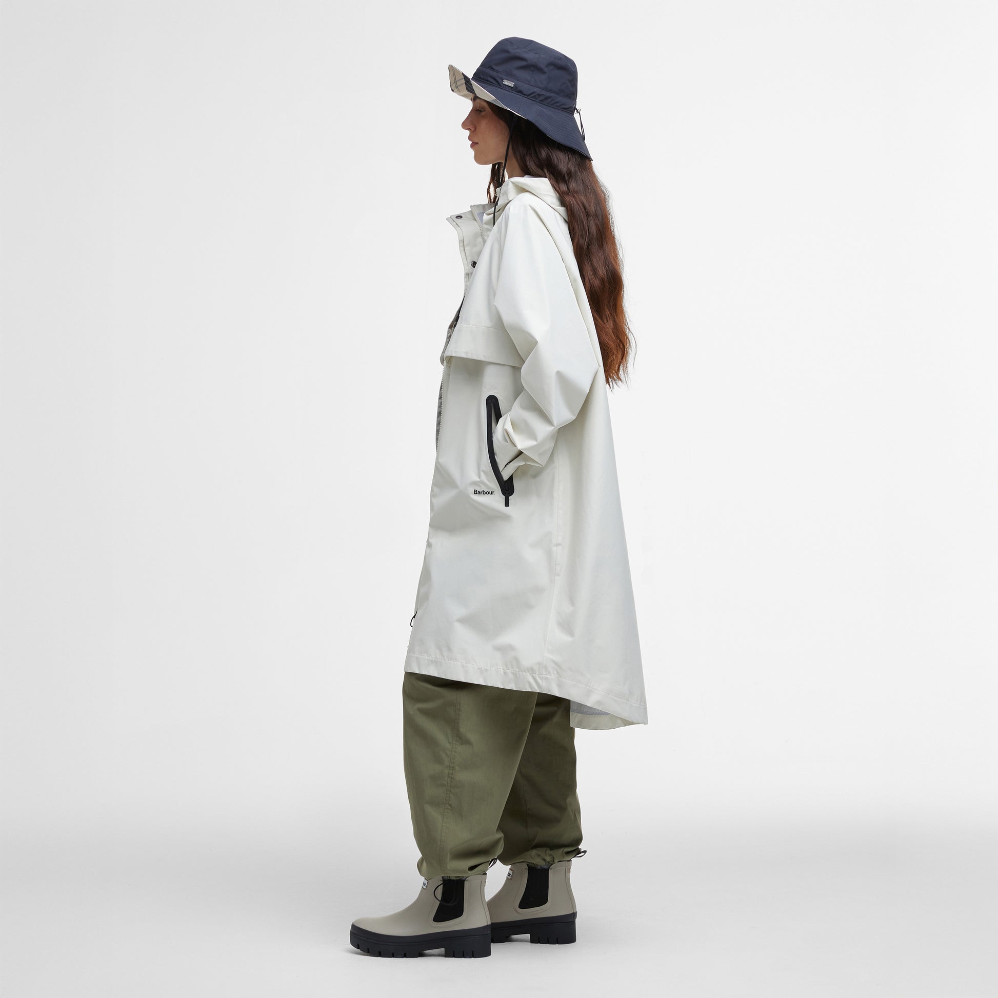 A person stands in profile against a white background, wearing the Barbour Jayla Waterproof Parka in white, paired with olive pants, beige boots with black soles, and a navy bucket hat—an ideal outfit for outdoor adventures.