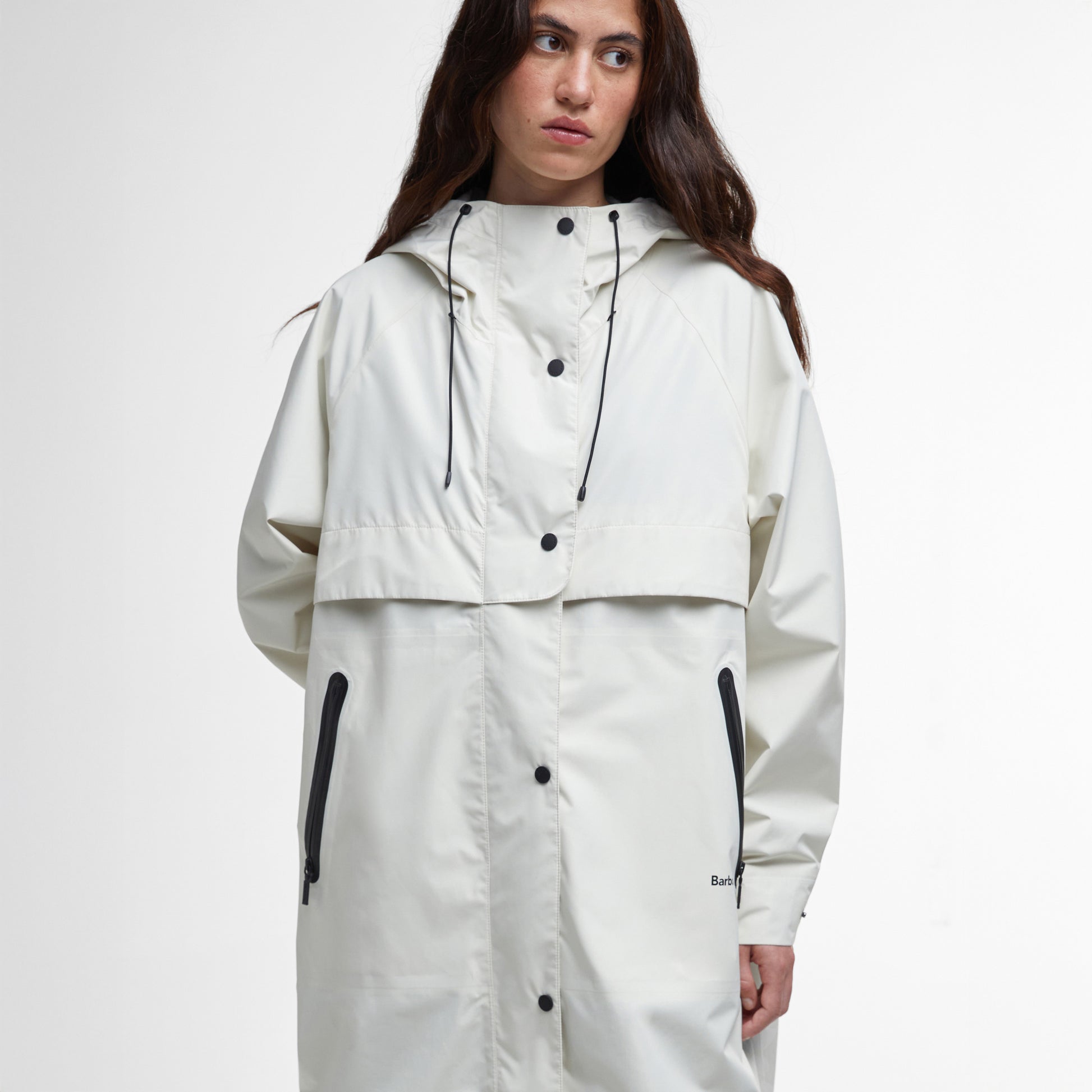 A person wears the Barbour Jayla Waterproof Parka—a long, off-white coat with black zippers and buttons—against a white background, showcasing its stylish design and weather-ready protection.