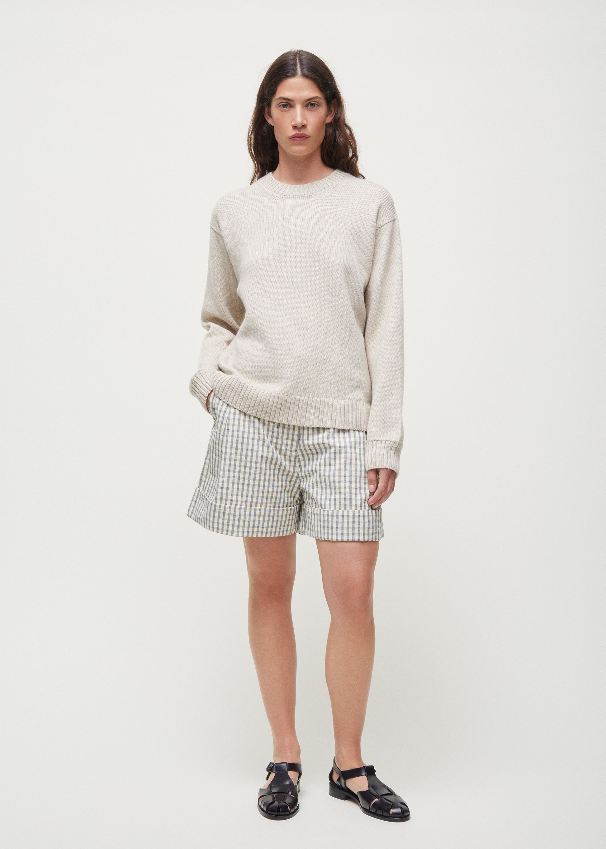 A person stands against a plain background wearing a light sweater, Aiayu's Lee Shorts Check made from organic Chetna cotton, and black shoes, with hands in pockets.