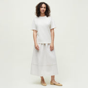 A woman with curly hair stands against a plain background, wearing a loose white short-sleeve shirt, the Aiayu Luka Seersucker Skirt—an ankle-length organic cotton design—and beige flat shoes.