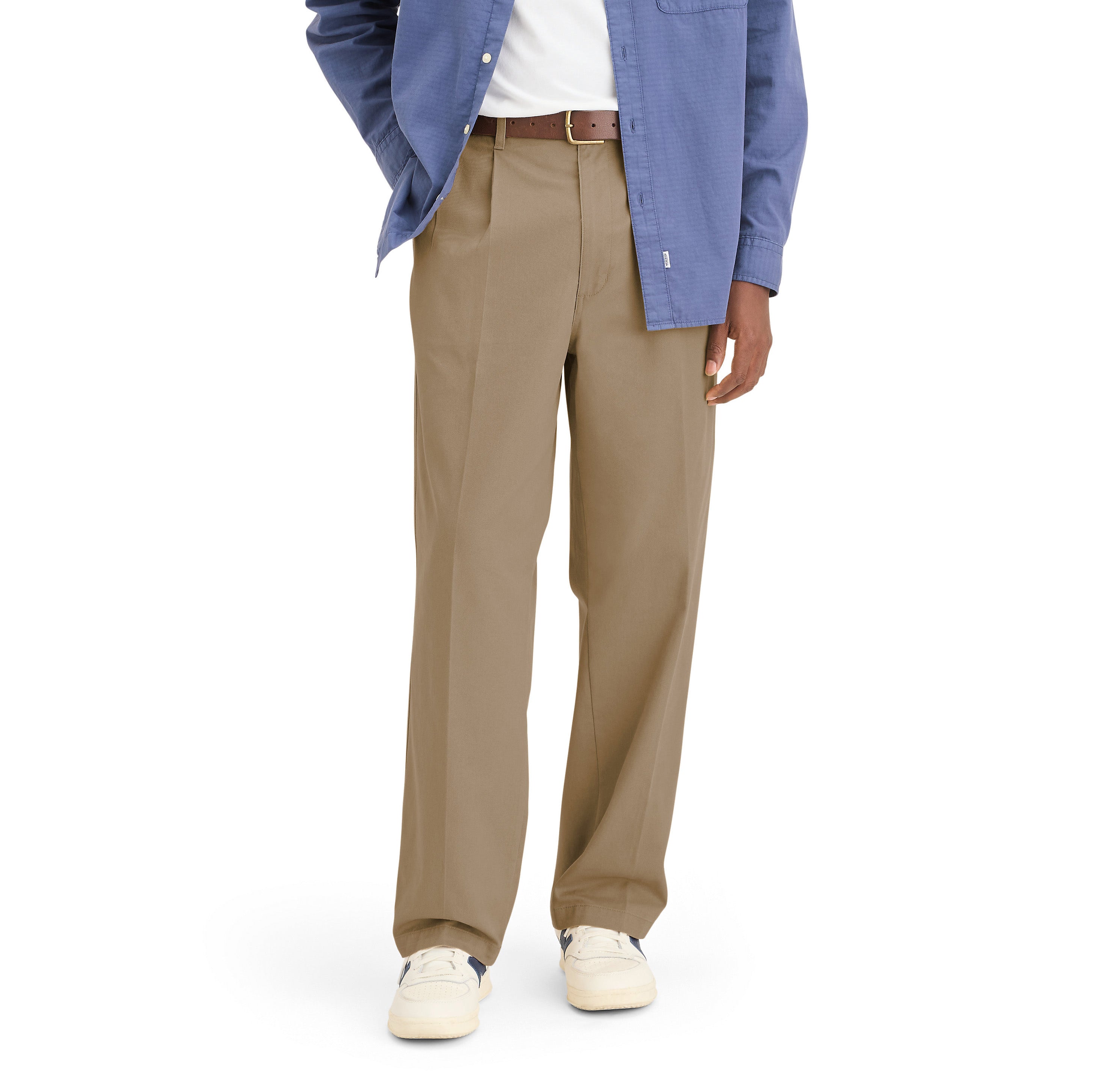 Wearing Dockers’ Genderless 86 Original Pleated Chino in tan, a white tee, blue button-up shirt, brown belt, and white sneakers, a person stands against a plain background. The chinos are made from recycled cotton.