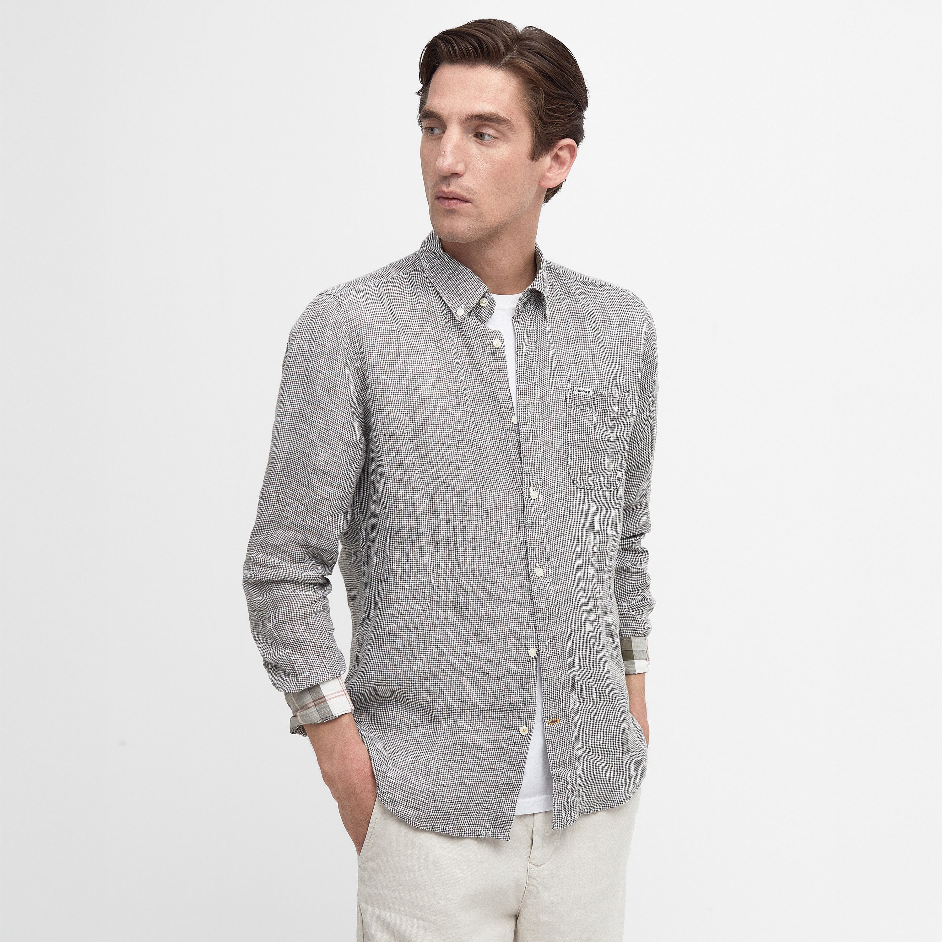 A person wears the gray Linton Tailored Shirt by Barbour, hands in pockets, gazing sideways against a white background. The shirt features a subtle micro houndstooth weave, adding understated elegance.