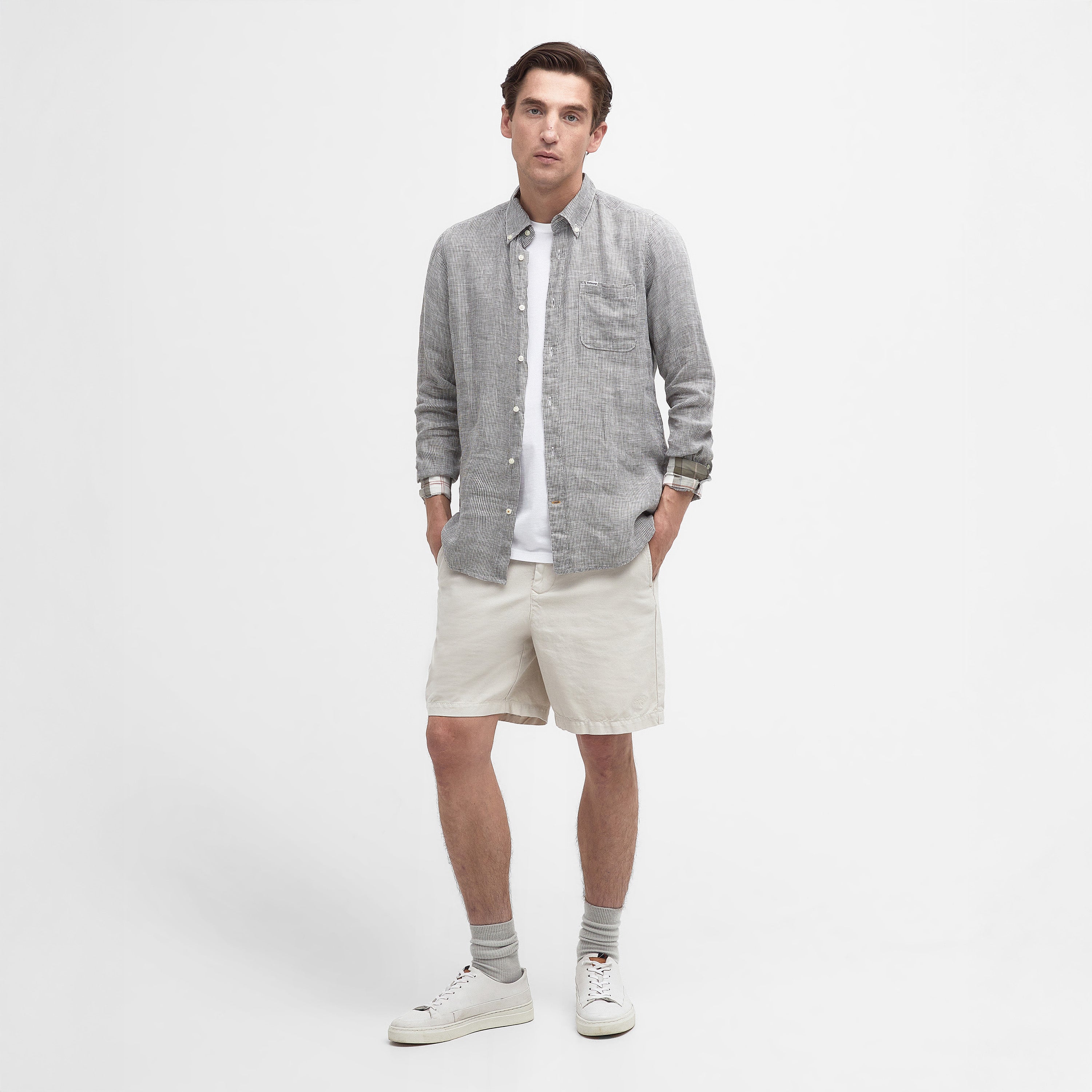 A person stands against a white background, wearing the Linton Tailored Shirt by Barbour over a white T-shirt, beige shorts, gray socks, and white sneakers.