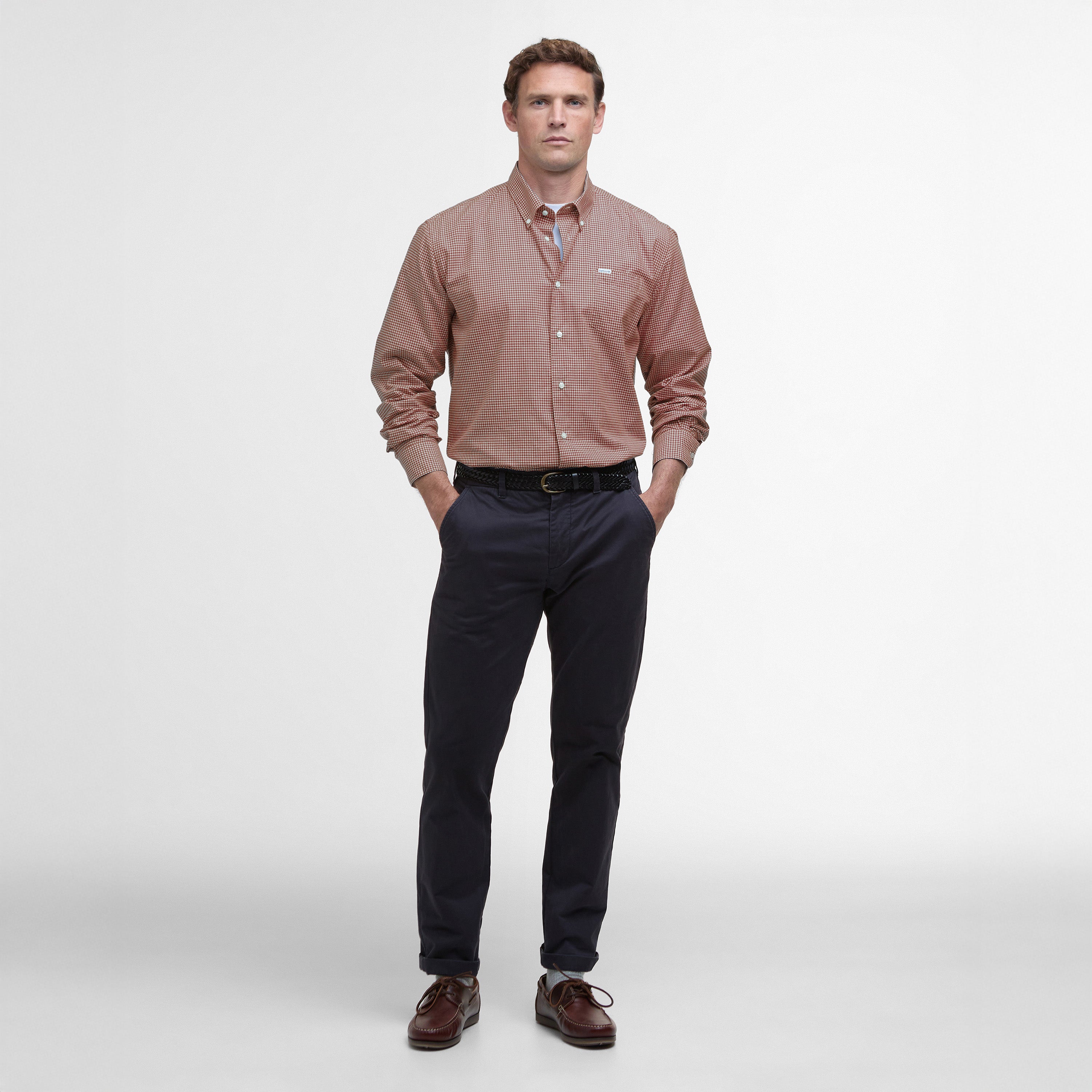 A man in a Barbour Finwell Gingham Tailored Fit Shirt, vibrant red and 100% cotton, pairs it with dark pants while standing against a plain white backdrop, hands casually in his pockets.