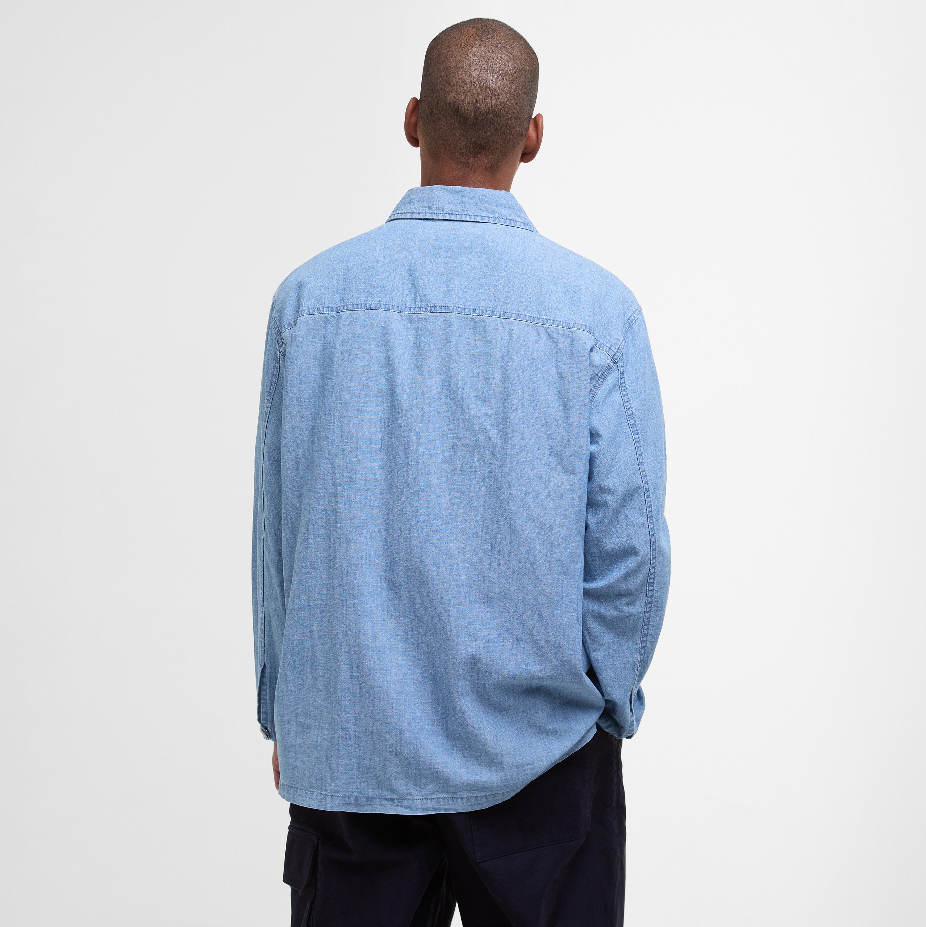 A person stands facing away from the camera, wearing the Barbour Markham Smock Shirt in light blue chambray and dark pants, set against a plain white background.