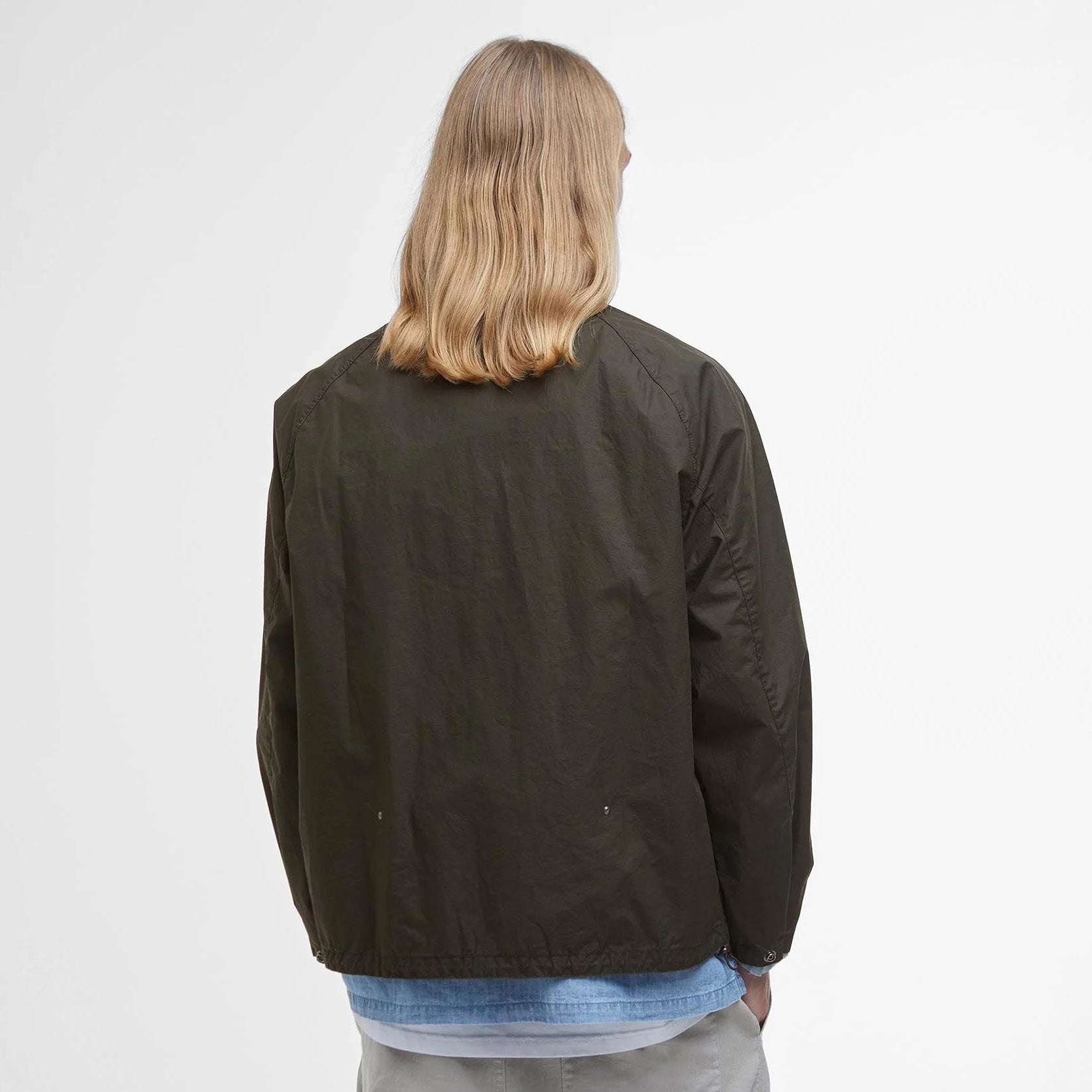A person with long blond hair, embracing countryside life, faces away wearing a Barbour Westmorland Showerproof Jacket over a blue shirt and light pants. The tranquil scene is enhanced by the simplicity of a plain white wall background.