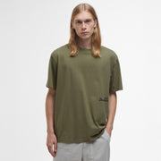A person with long blond hair stands against a white background, wearing an oversized olive green Regence Graphic T-Shirt by Barbour and light gray pants, exuding an effortlessly chic vibe.