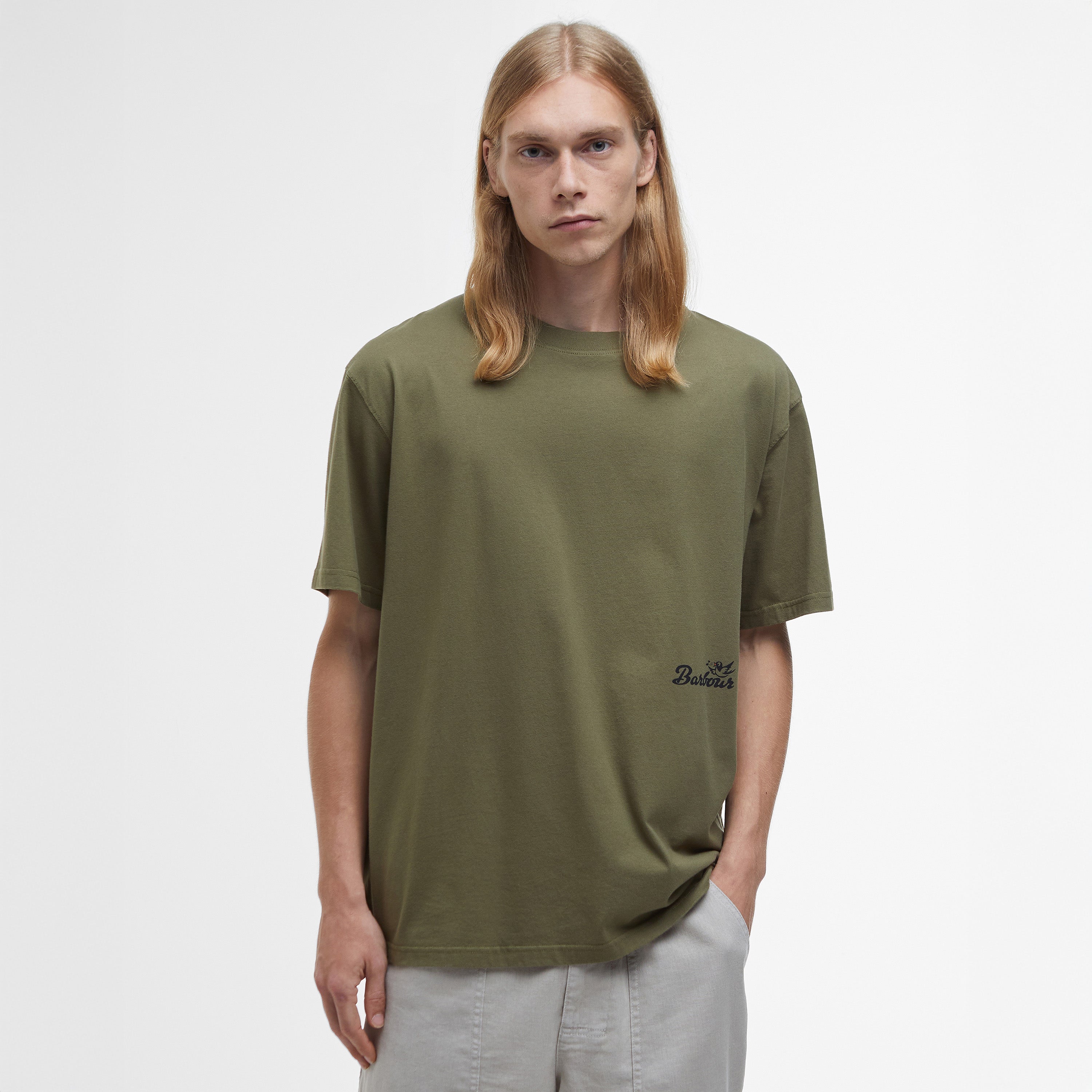 A person with long blond hair stands against a white background, wearing an oversized olive green Regence Graphic T-Shirt by Barbour and light gray pants, exuding an effortlessly chic vibe.