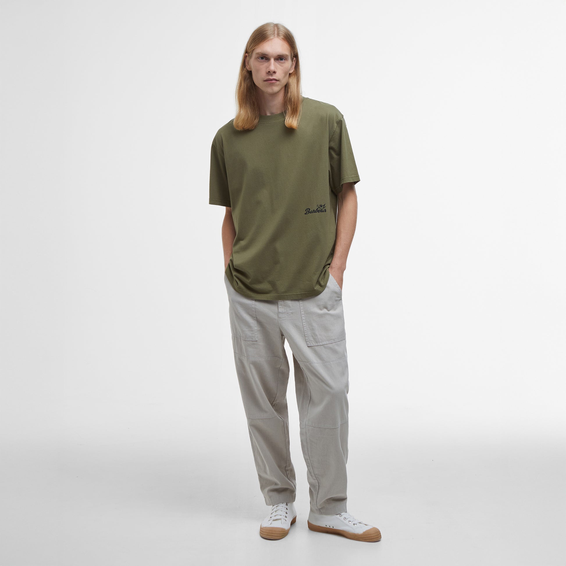 A person with long blonde hair stands wearing an oversized Regence Graphic T-Shirt by Barbour, light gray pants, and white sneakers against a plain background.