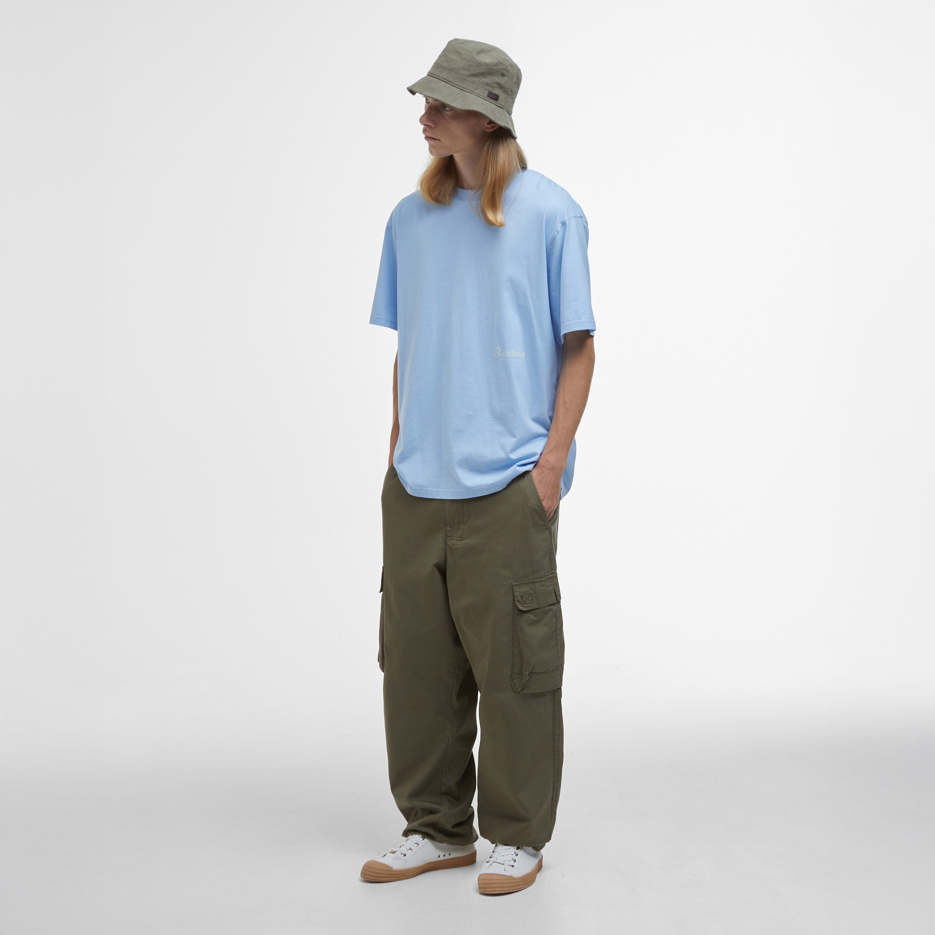 Wearing a Barbour Newclyde Graphic T-Shirt and a bucket hat, a person stylishly complements their green cargo pants and white sneakers against a plain white background.