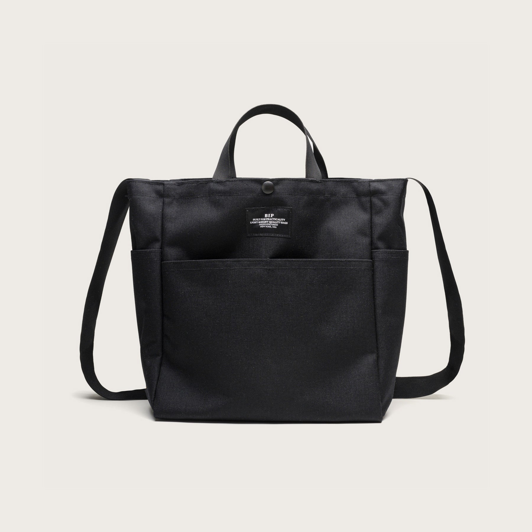 The Multi Pocket Bag Medium by Bags in Progress is a black, medium-sized water-repellent nylon shoulder bag with short handles, a long strap, front pockets, and an upper front label, shown against a plain light background.