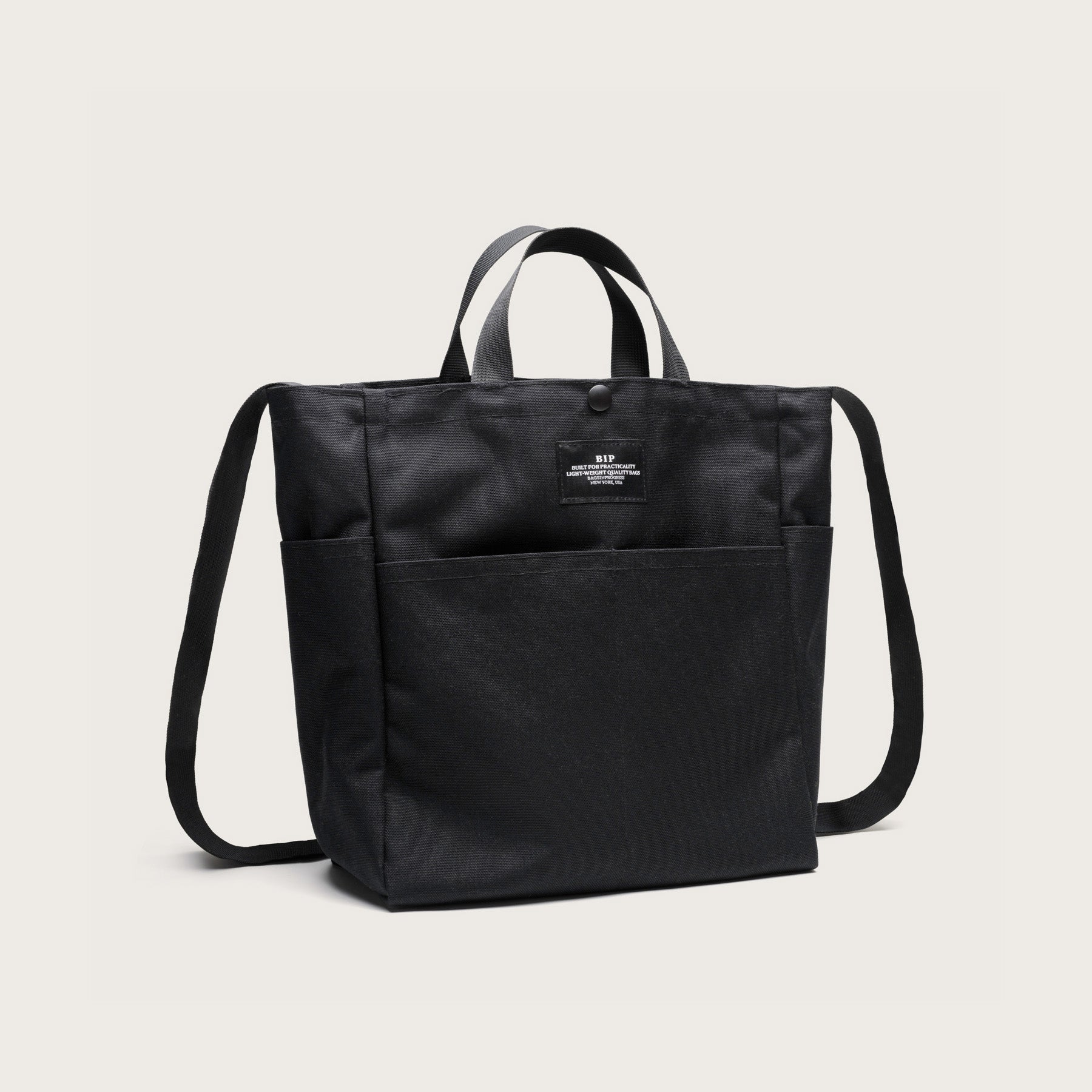 The Bags in Progress Multi Pocket Bag Medium is a black nylon canvas tote with short handles, long shoulder straps, front pockets, and a small front label. Water repellent and practical, it’s shown against a plain light background.