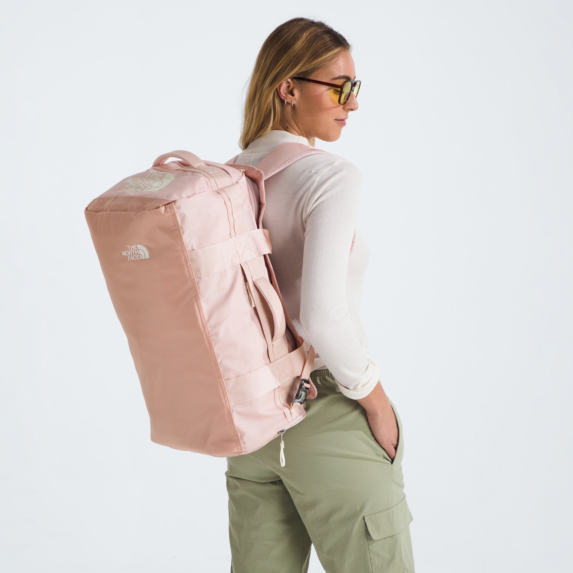 A woman in sunglasses and casual clothing carries a large, light pink The North Face Base Camp Voyager Duffel - 42L over one shoulder, showcasing its lightweight design against a plain white background.