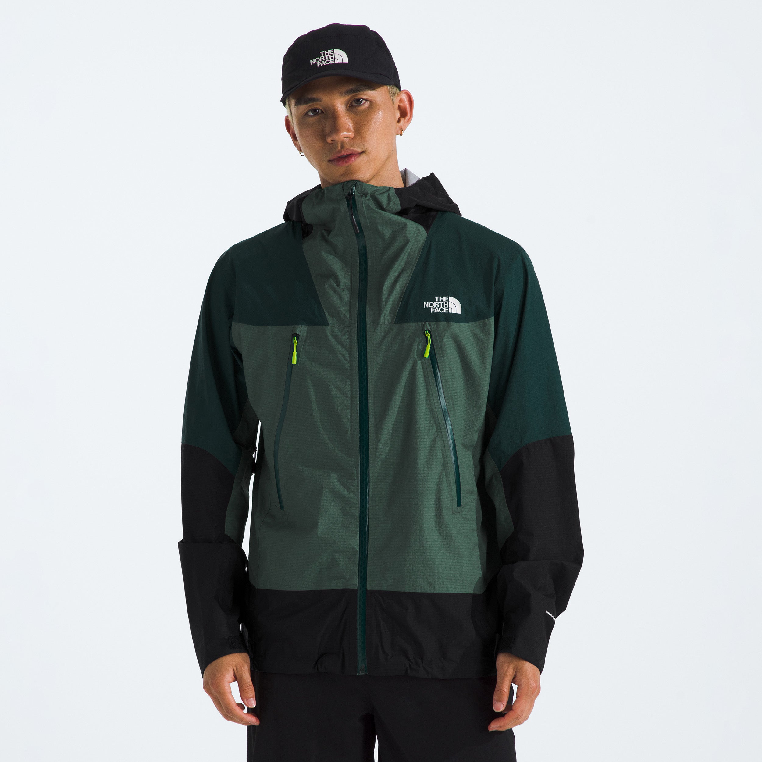 A person wearing The North Face's M Signal 2.5L DryVent jacket in dark green and black, complete with a hood, and a matching cap stands against a plain background.