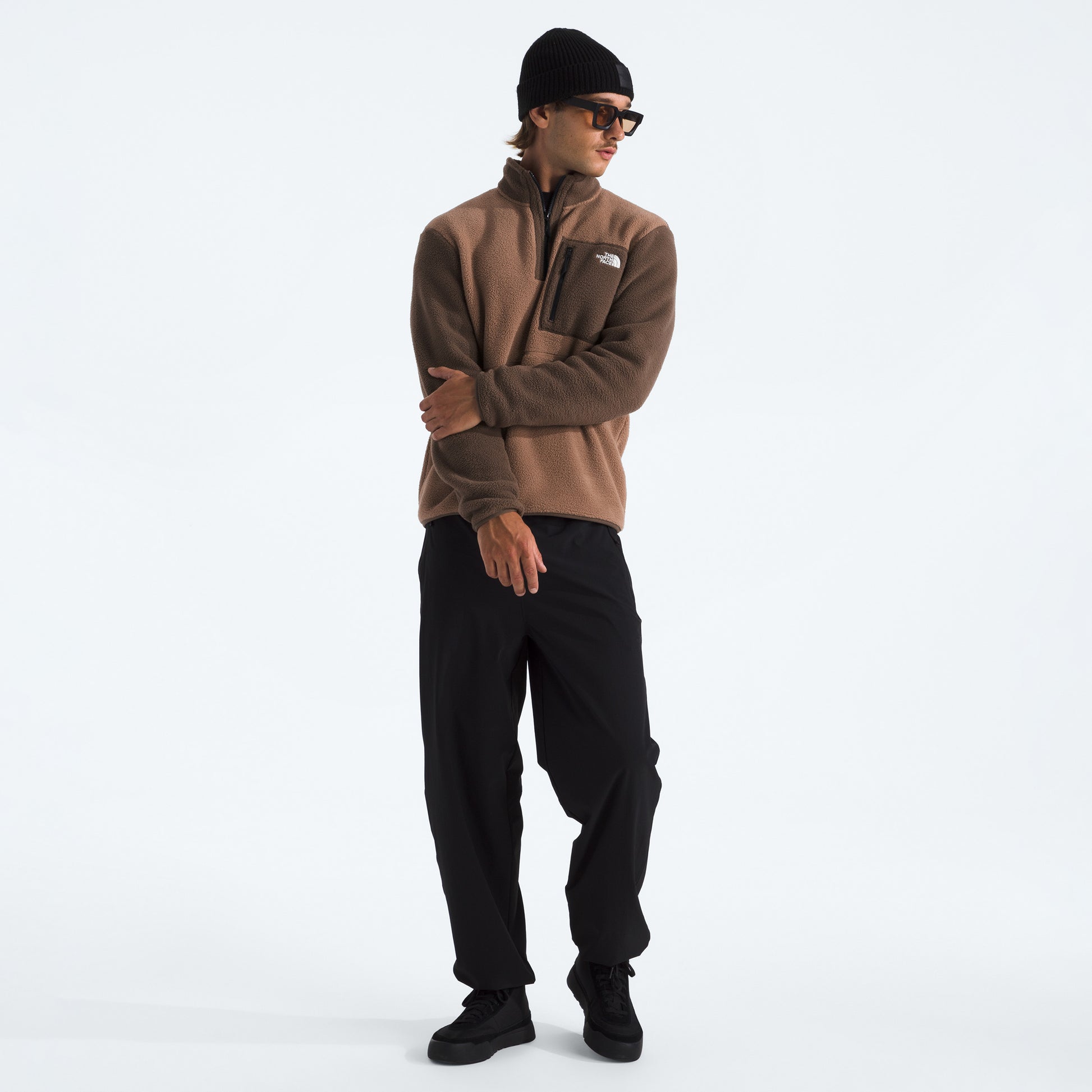 A person wears The North Face M Yumiori 1/4 Zip recycled fleece jacket, black pants, a black beanie, and sunglasses while standing against a plain white background.