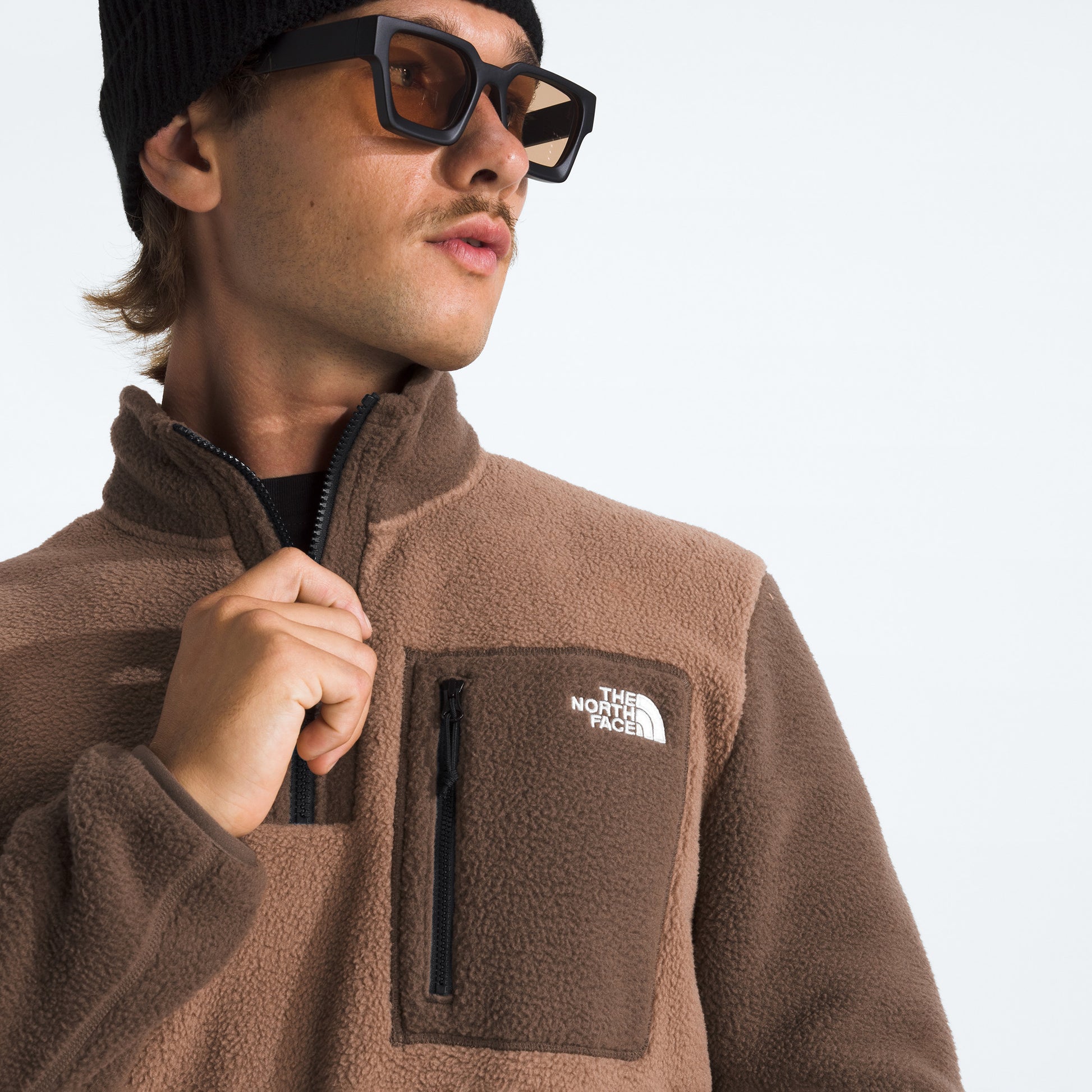 A person in sunglasses, a black beanie, and a brown The North Face M Yumiori 1/4 Zip fleece jacket with recycled material and a zippered chest pocket looks to the side.