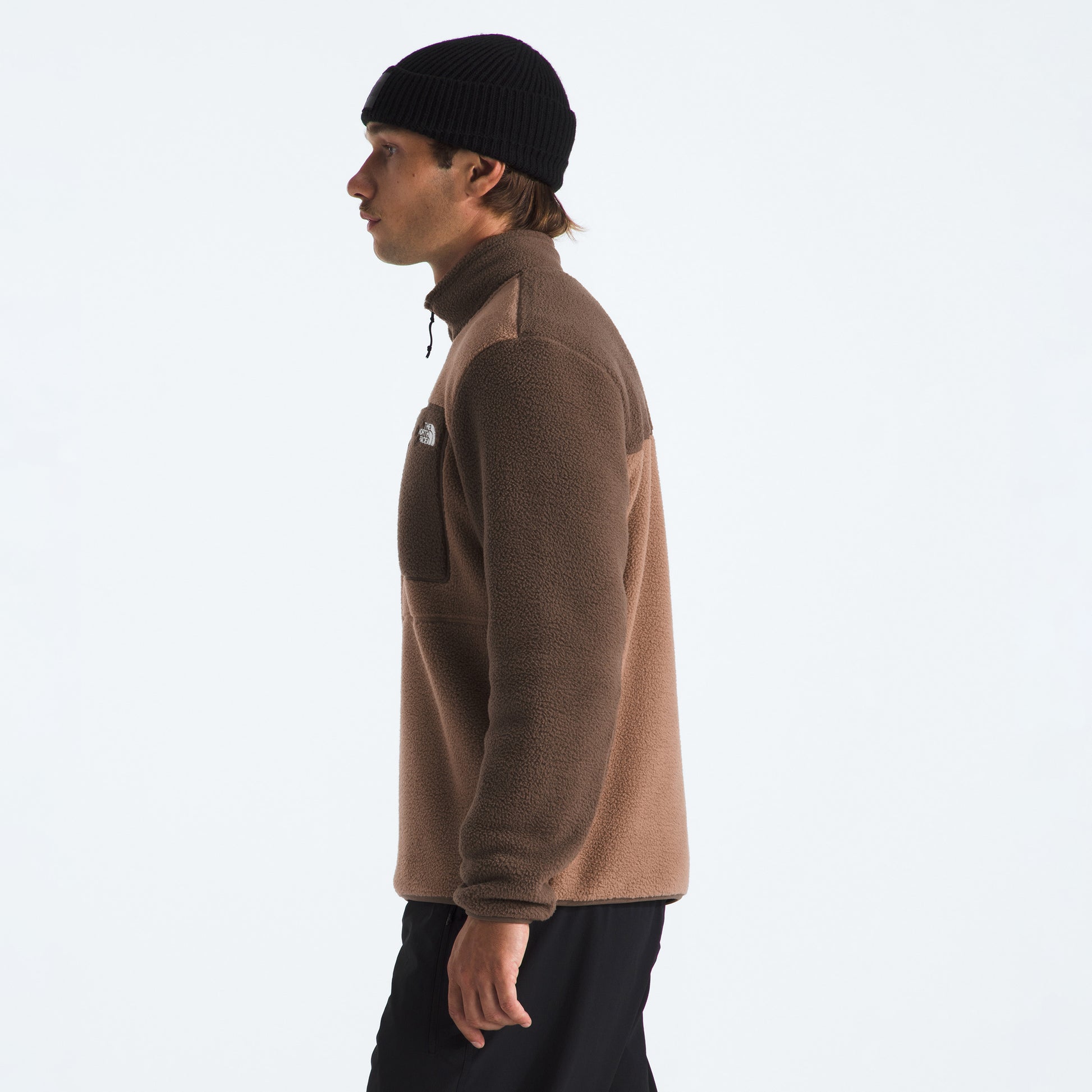A person stands in profile against a plain light background, wearing The North Face M Yumiori 1/4 Zip recycled fleece jacket in brown, black pants, and a black knit beanie.