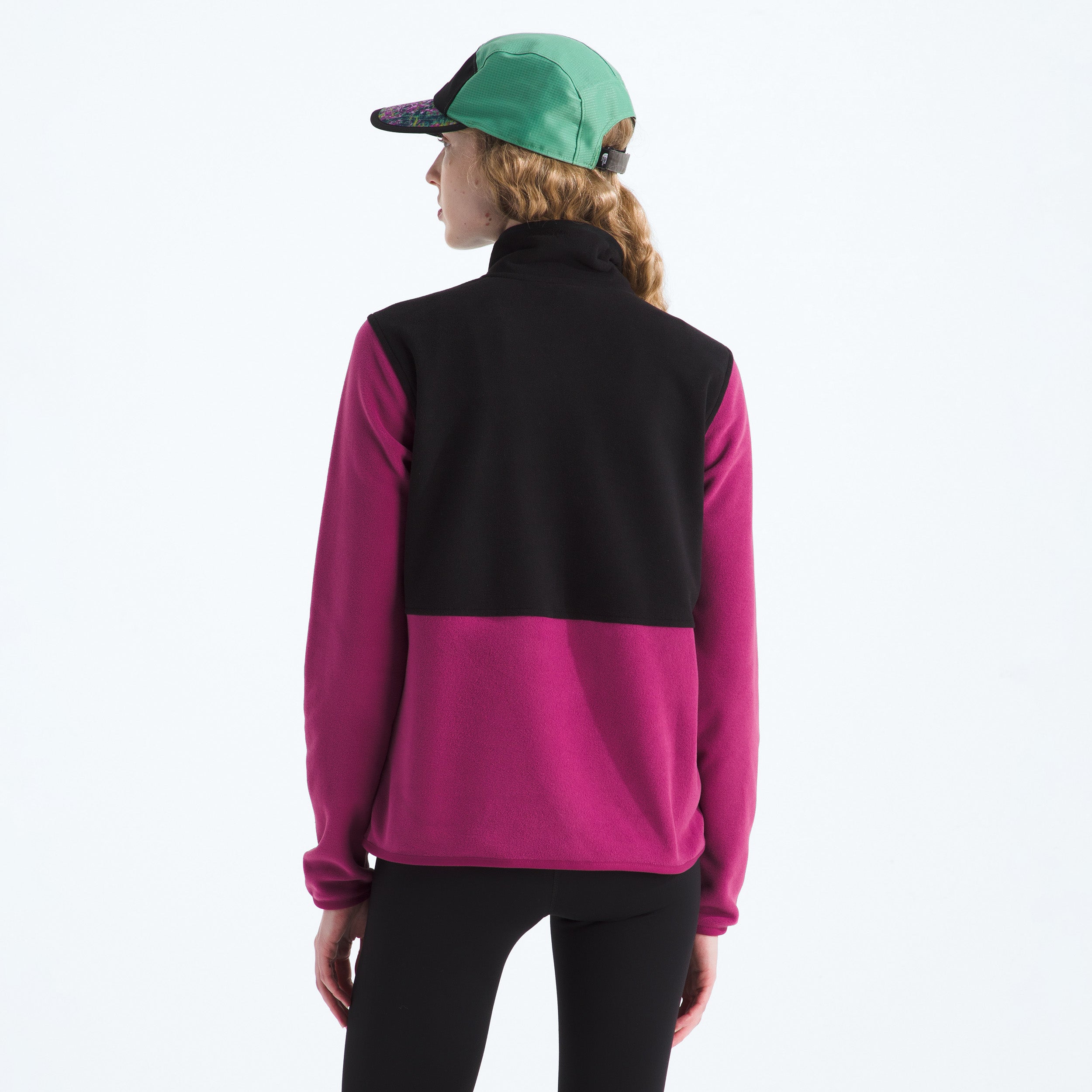 The person is wearing a W Glacier Fleece Jacket in black and magenta by The North Face, paired with black leggings and a green-and-black cap. The jacket's lightweight fleece provides style and comfort from behind and incorporates sustainability through recycled fabric.