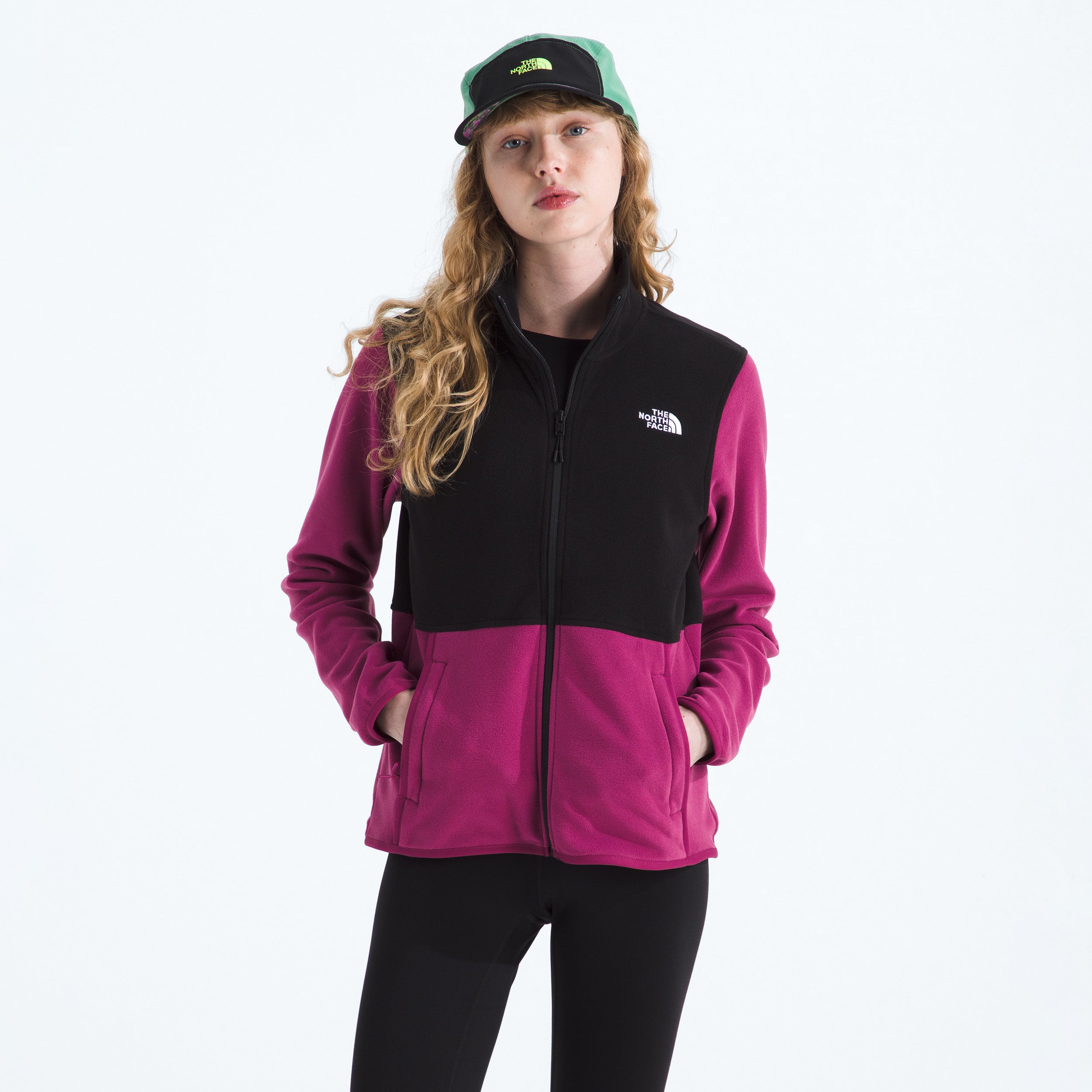 A person wears The North Face W Glacier Fleece Jacket in black and magenta, made from recycled fabric, with a green and black cap, standing hands in pockets against a plain background.