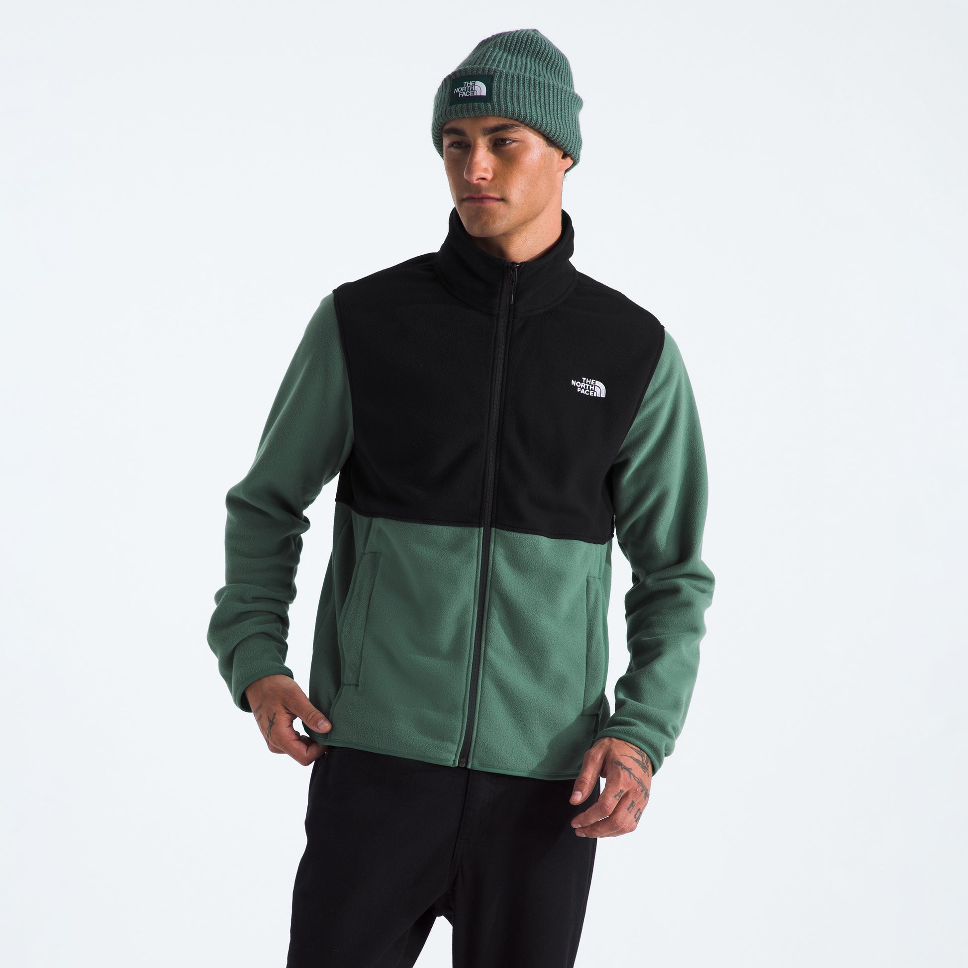 A person wearing The North Face M Glacier Fleece Jacket in green and black, with black pants and a green knit beanie, stands against a plain white background.