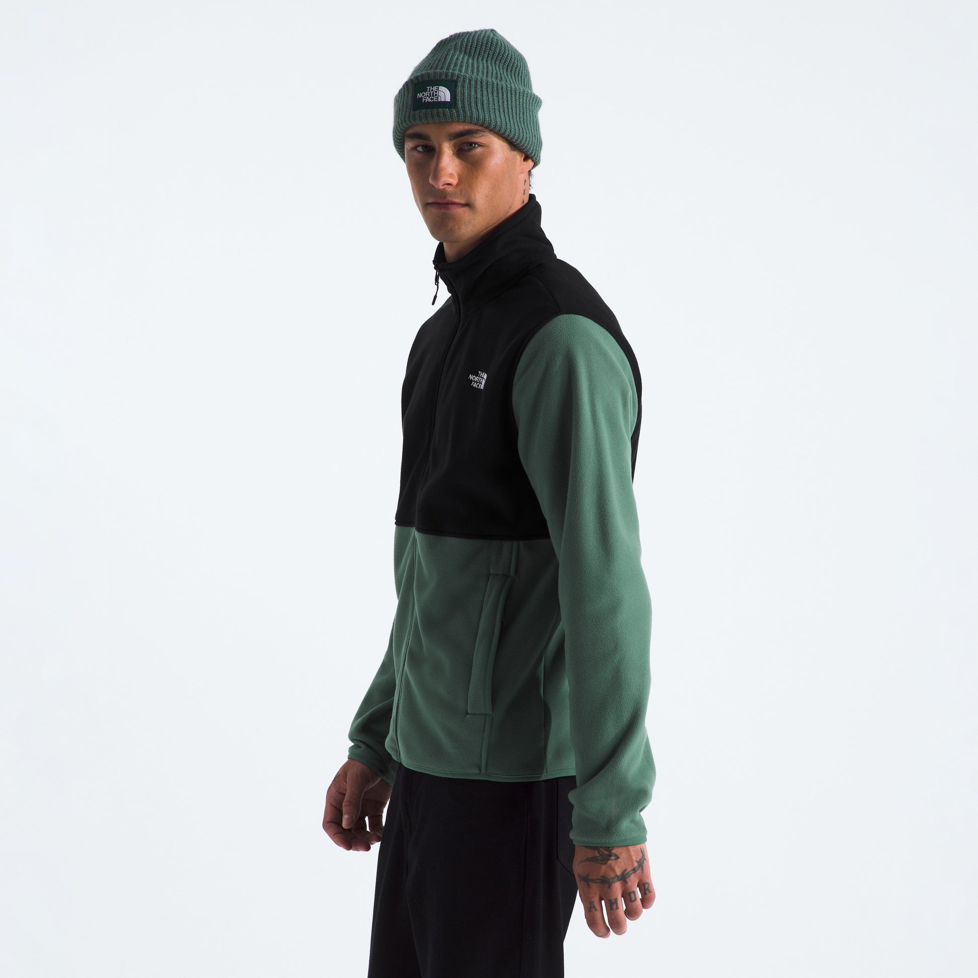 A person wears a green beanie, black pants, and The North Face M Glacier Fleece Jacket—made from recycled fabric—while standing against a plain white background.