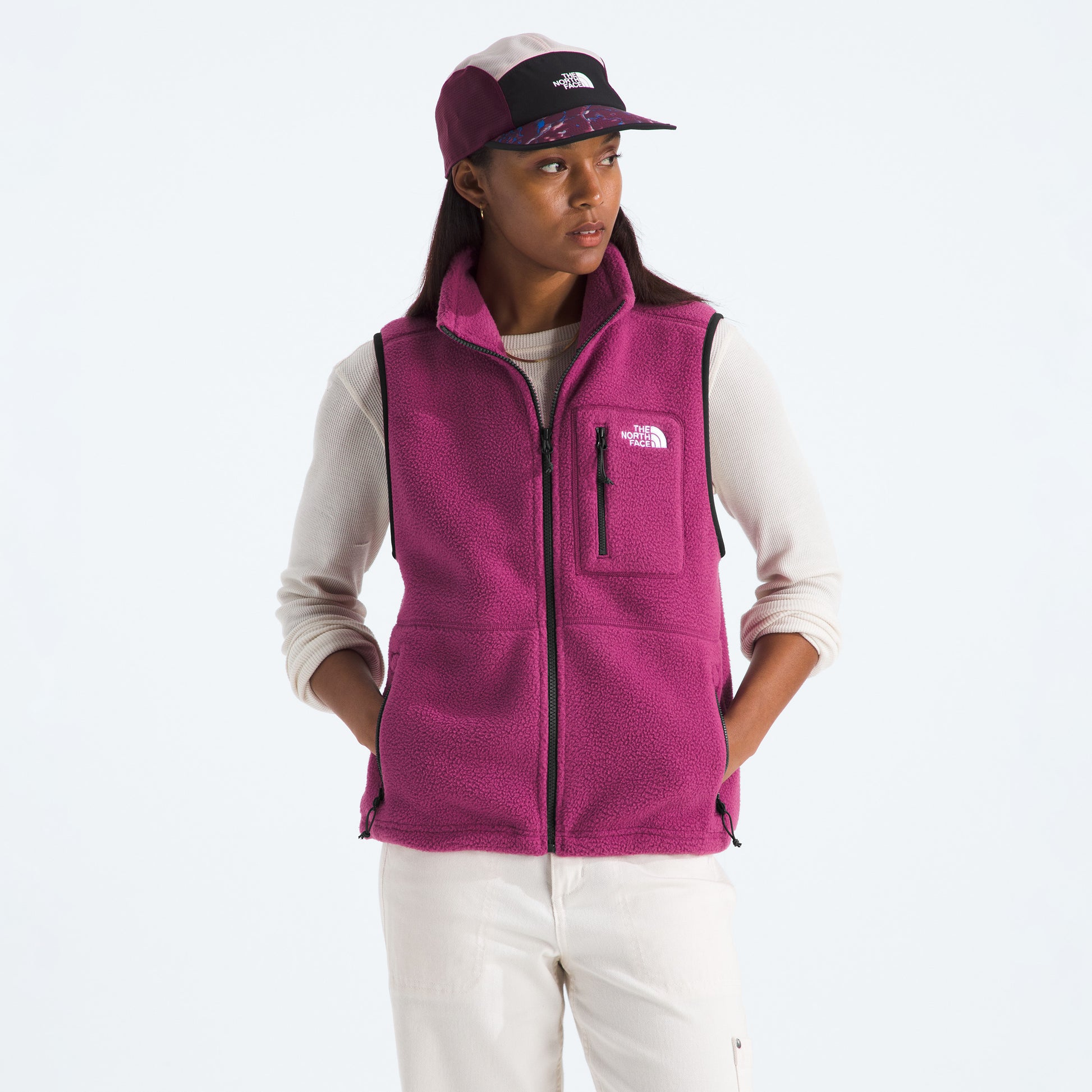 A person wears The North Face W Yumiori Vest in magenta with an adjustable hem, paired with an off-white long-sleeve shirt, off-white pants, and a two-tone cap, standing against a plain background.
