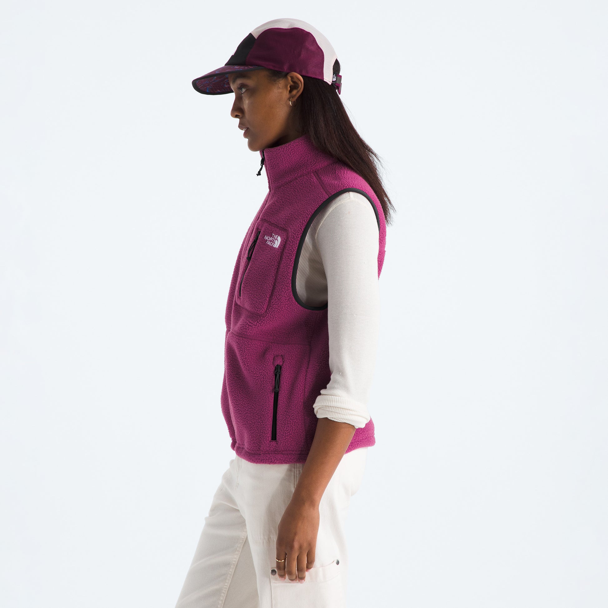 A person in profile wears a magenta The North Face W Yumiori Vest with adjustable hem, paired with a white long-sleeve shirt, white pants, and a magenta cap, standing against a plain light background.