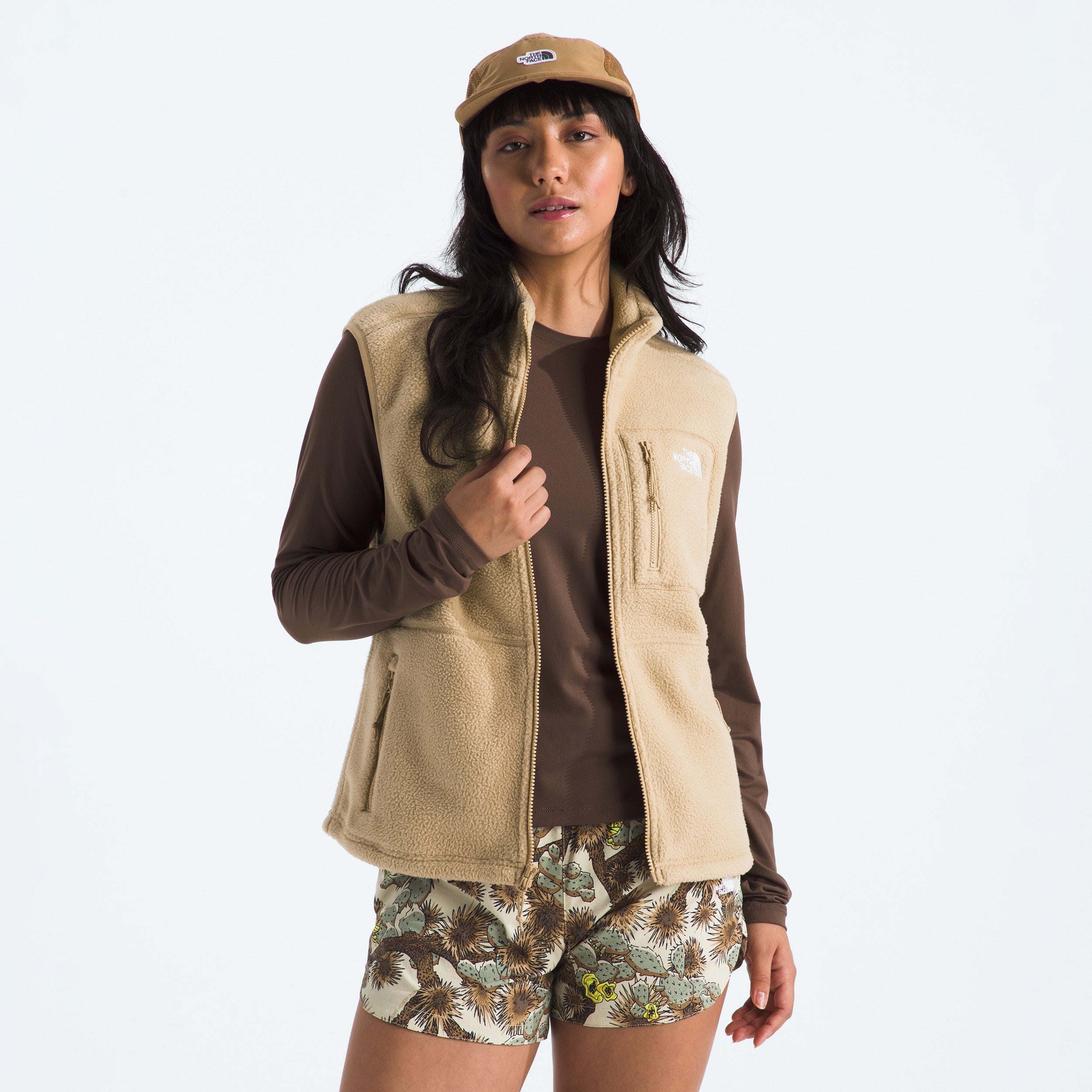 Dressed in The North Face's W Yumiori Vest crafted from high-pile fleece, a person stands against a plain backdrop, complemented by a brown long-sleeve shirt, floral shorts, and a tan cap.