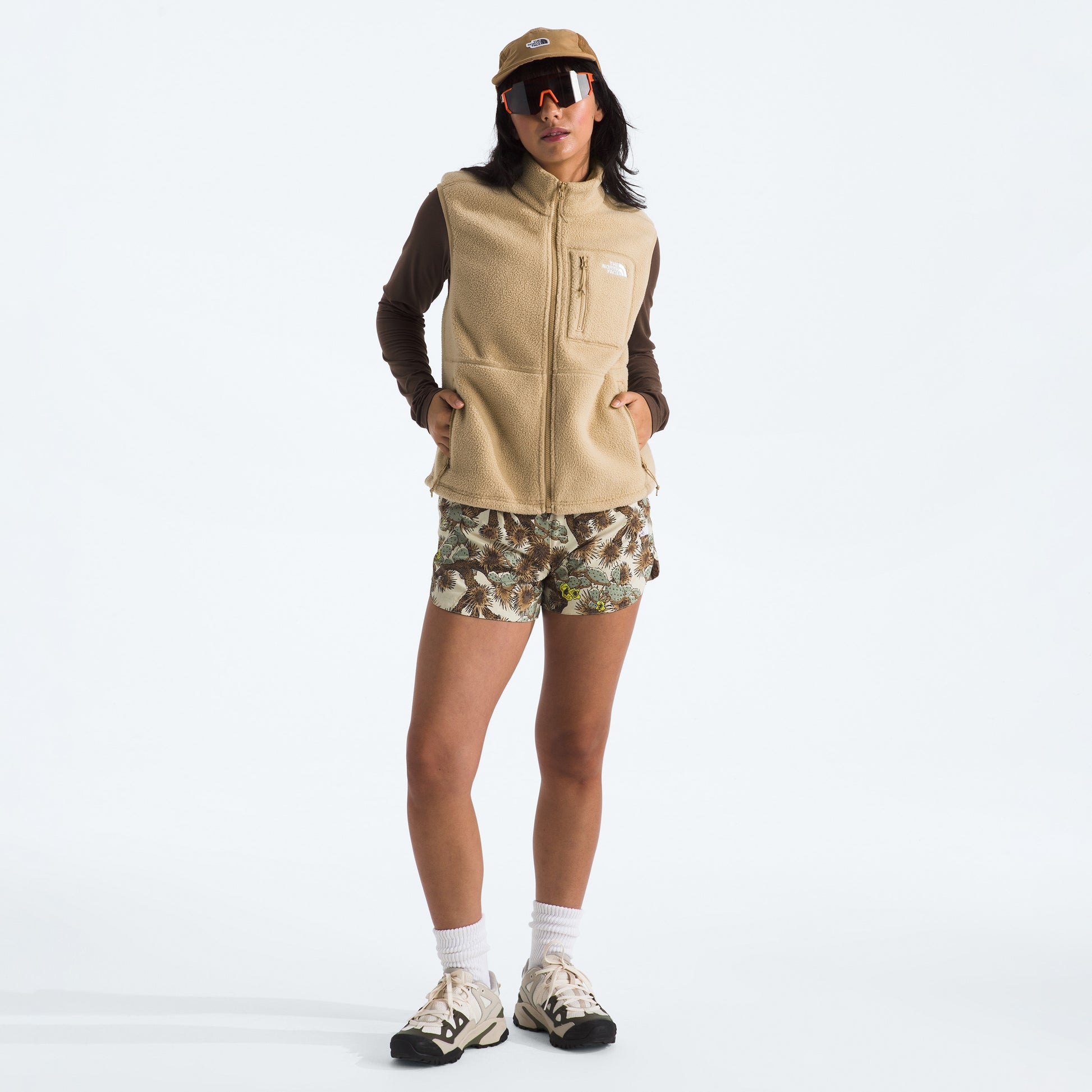 A person wearing The North Face W Yumiori Vest with a brown long-sleeve shirt, camouflage shorts, white socks, sneakers, a brown cap, and large sunglasses stands against a plain white background.