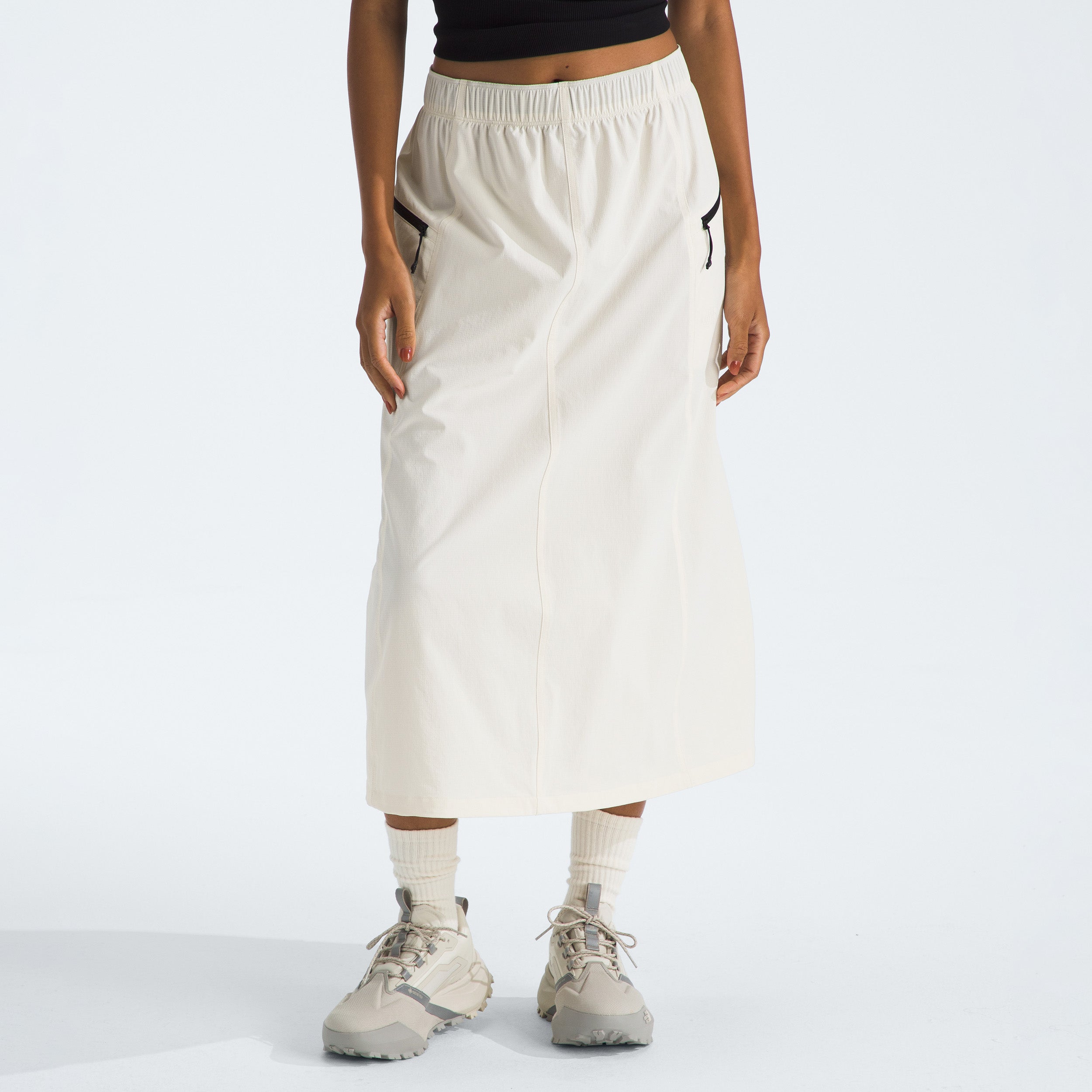 A person in a black top and gray sneakers stands against a plain white background wearing The North Face's W North Dome Wind Skirt, which is long and white with black pockets.