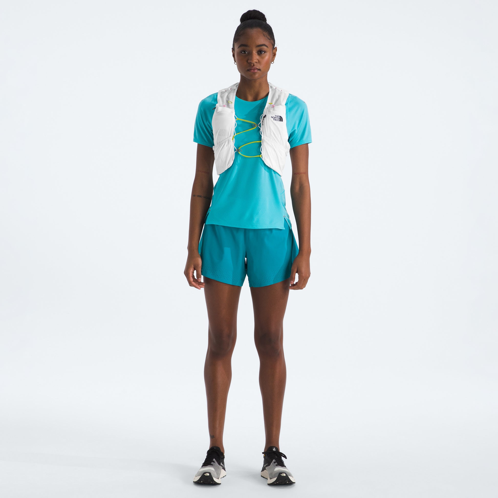 A person in a turquoise athletic outfit and black sneakers stands against a white background, wearing The North Face U Summit Run Vest 5—ideal for race day.