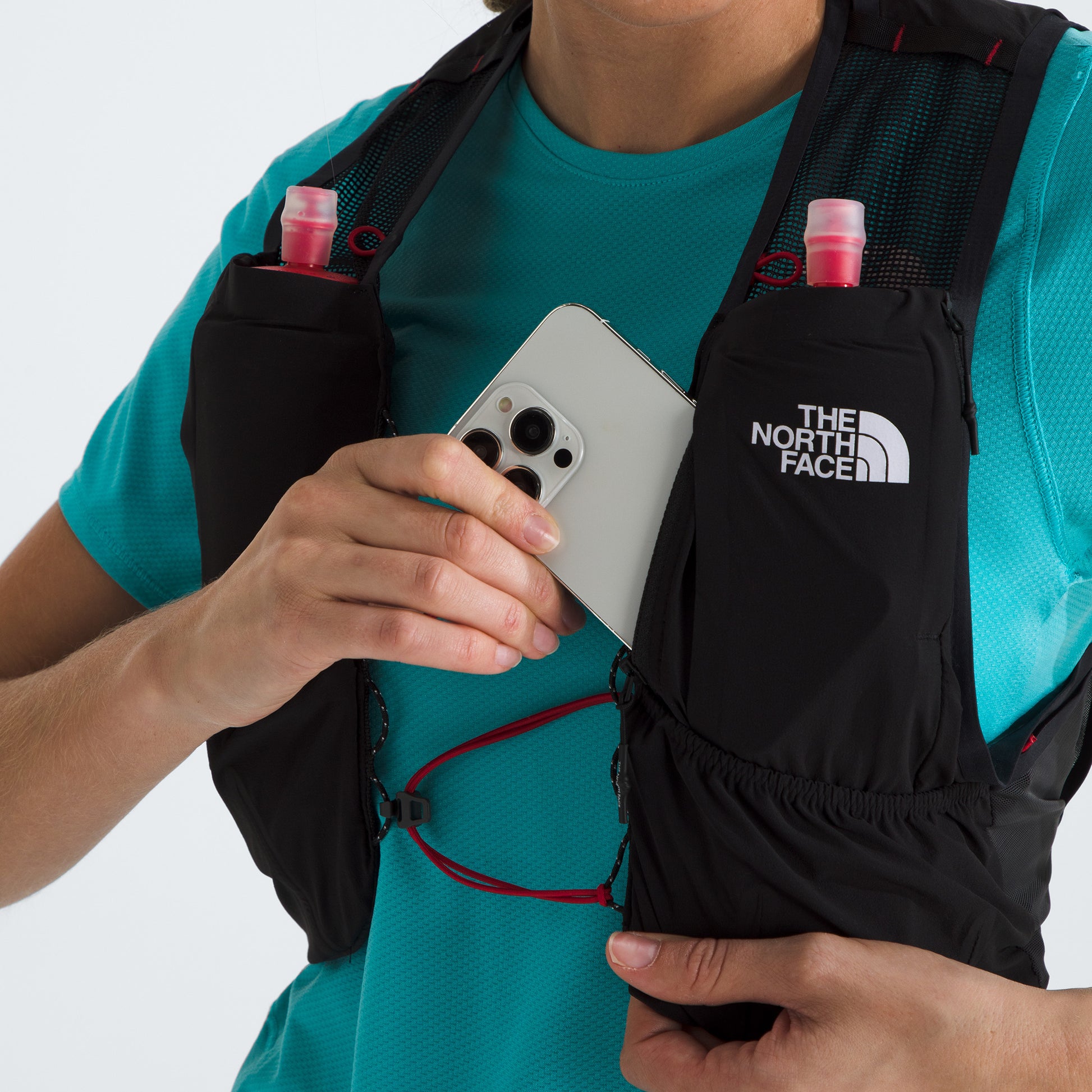 The person in a teal shirt is placing a smartphone into their black U Summit Run Vest 5 by The North Face, with two pink bottles visible in other pockets as they gear up for race day.